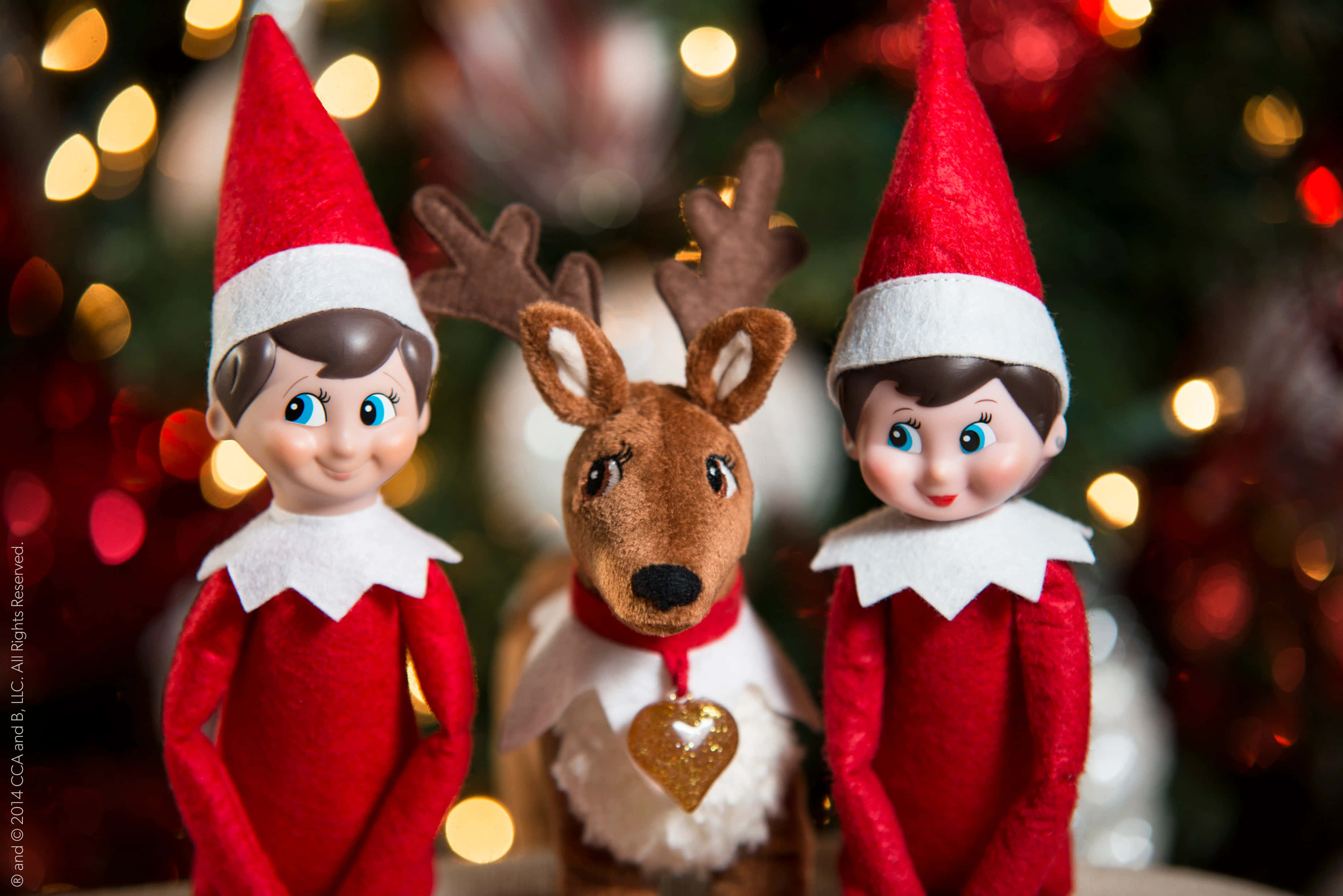 Christmas Elf 桌布 壁紙 — 兩個微笑的精靈娃娃夾著一個帶閃爍心形標籤的毛絨馴鹿，背景是散景聖誕樹。