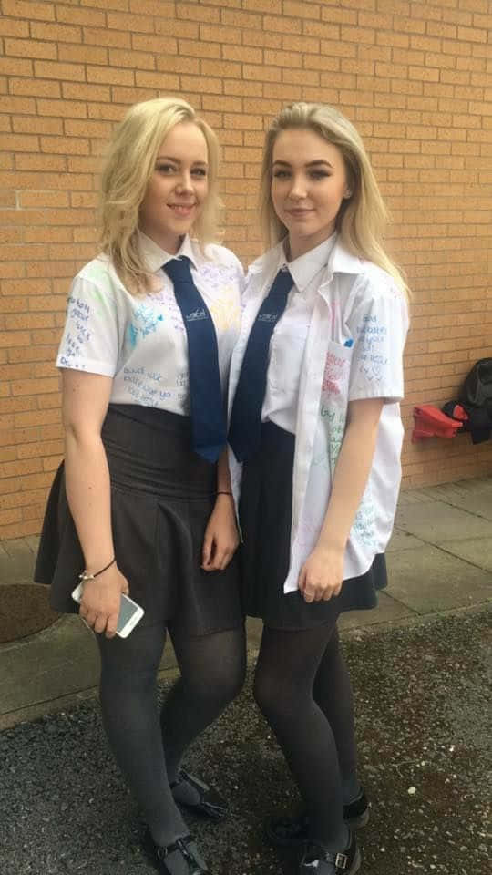 美學 桌布 壁紙 — 兩個微笑的金髮女學生穿著制服，緊靠著磚牆拿著手機。