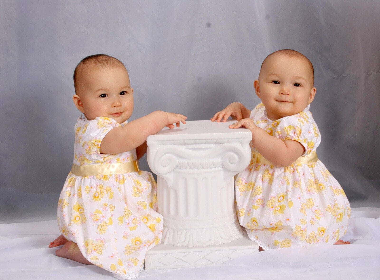 Cute Babies 桌布 壁紙 — 兩個微笑的雙胞胎嬰兒穿著相配的花裙，跪在白色古典柱子的兩側，背景是柔和的灰色。