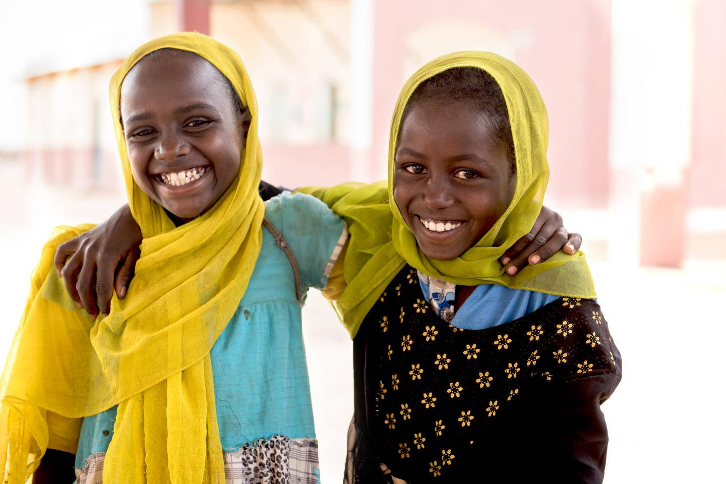 Sudan 桌布 壁紙 — 兩個快樂的女孩戴著鮮黃色頭巾，在柔和的粉紅色建築背景下互相擁抱，溫暖地微笑。