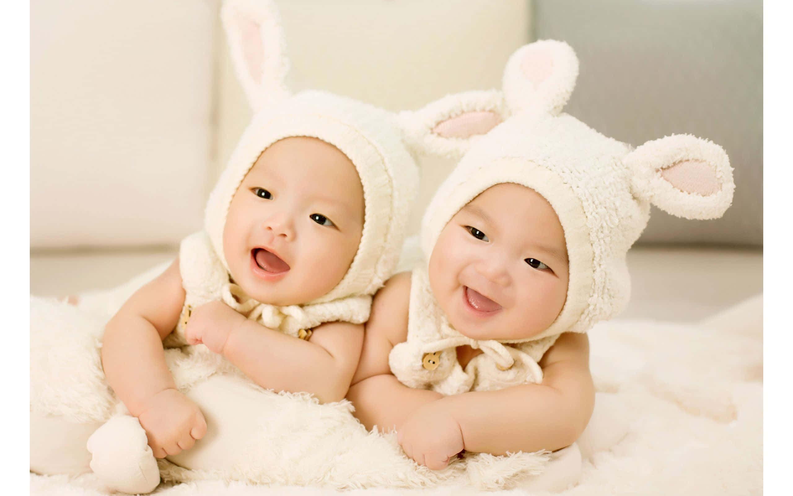 twins 桌布 壁紙 — 兩個快樂的嬰兒戴著毛茸茸的白色兔帽，坐在柔軟的白色地毯上，露出燦爛的笑容。