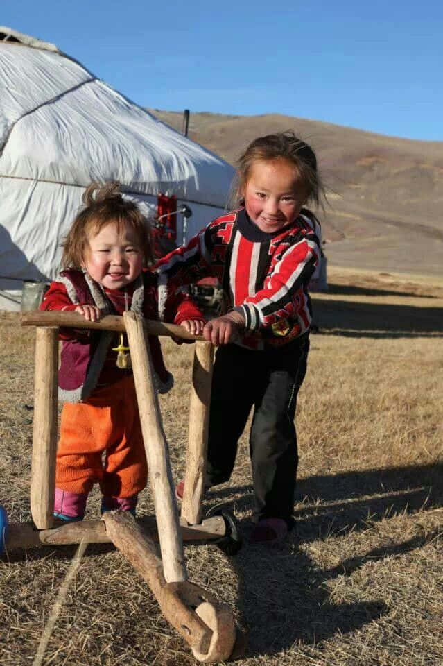 Mongolian 桌布 壁紙 — 兩個快樂的孩子在乾燥的草地上，在晴朗的藍天下，在蒙古包前推著木頭車。