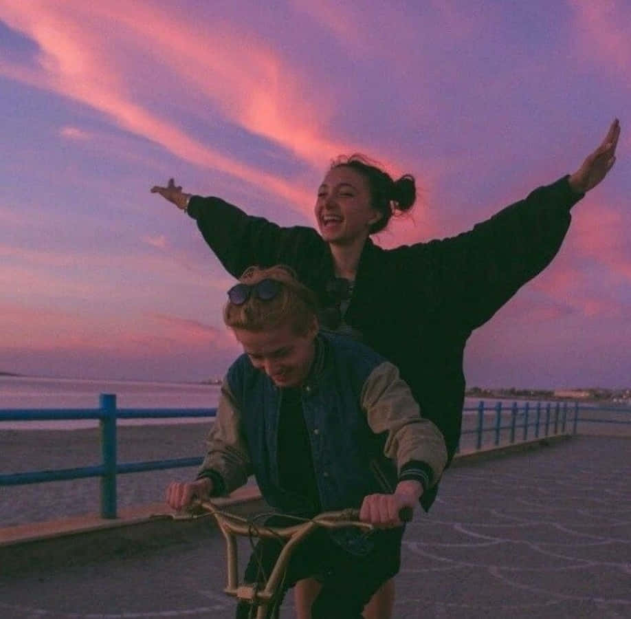Falling In Love 桌布 壁紙 — 兩個快樂的朋友在充滿活力的粉紅色和紫色日落天空下沿著海濱長廊騎自行車。