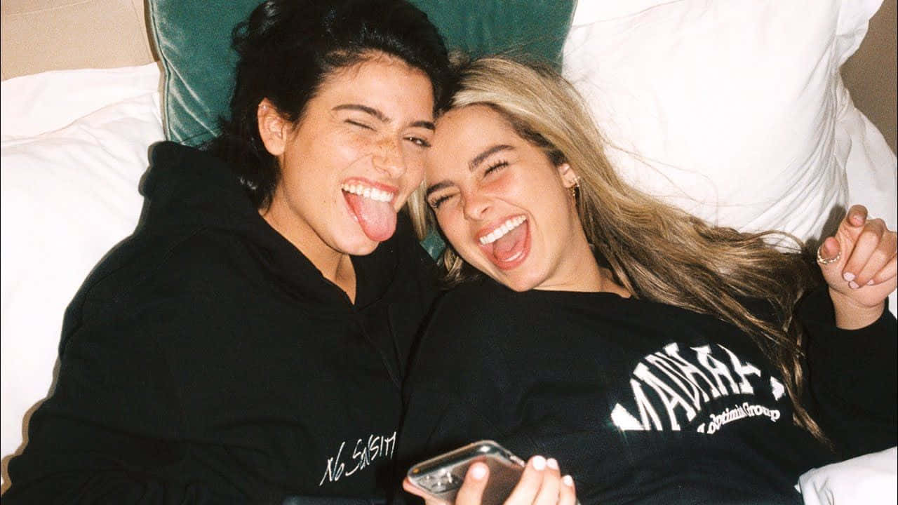 Charli And Addison 桌布 壁紙 — 兩個快樂的朋友躺在床上，白色枕頭和綠色床頭板，笑著伸出舌頭。