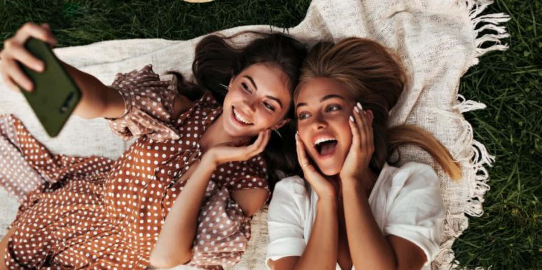 Friends 桌布 壁紙 — 兩個快樂的朋友躺在草地上的一條流蘇毛毯上，一邊笑著一邊用綠色手機自拍。