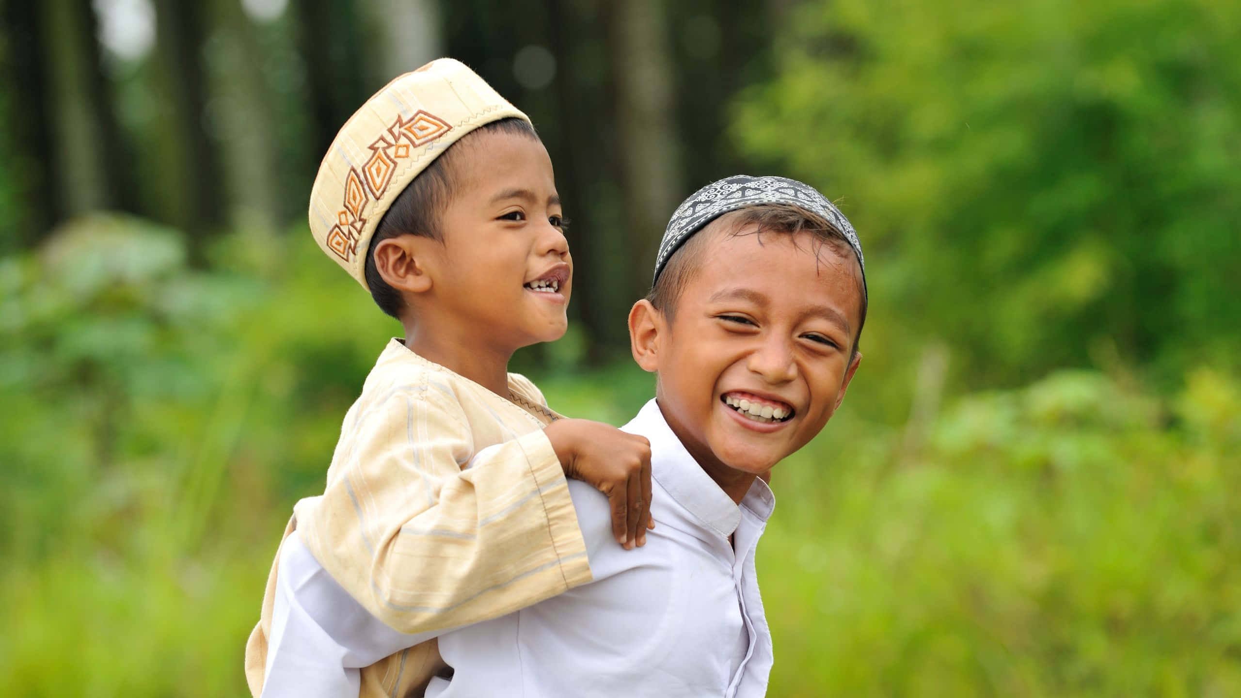 muslim 桌布 壁紙 — 兩個快樂的男孩戴著傳統帽子，在茂密的綠色森林中玩騎馬打仗。