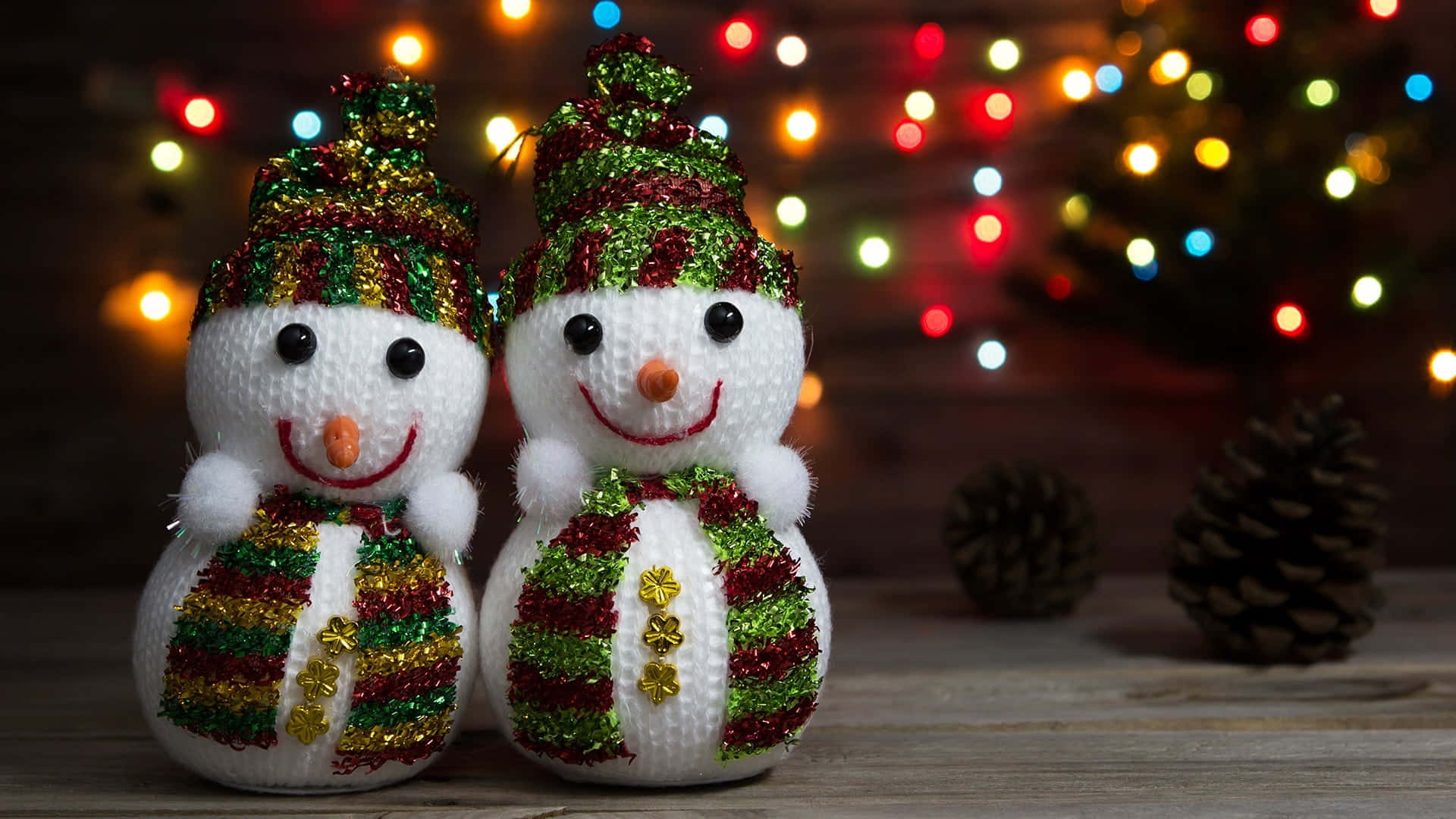 Couples Christmas 桌布 壁紙 — 兩個快樂的編織雪人，戴著閃亮的帽子和圍巾，站在木質表面上，背景是色彩繽紛的聖誕燈飾散景。