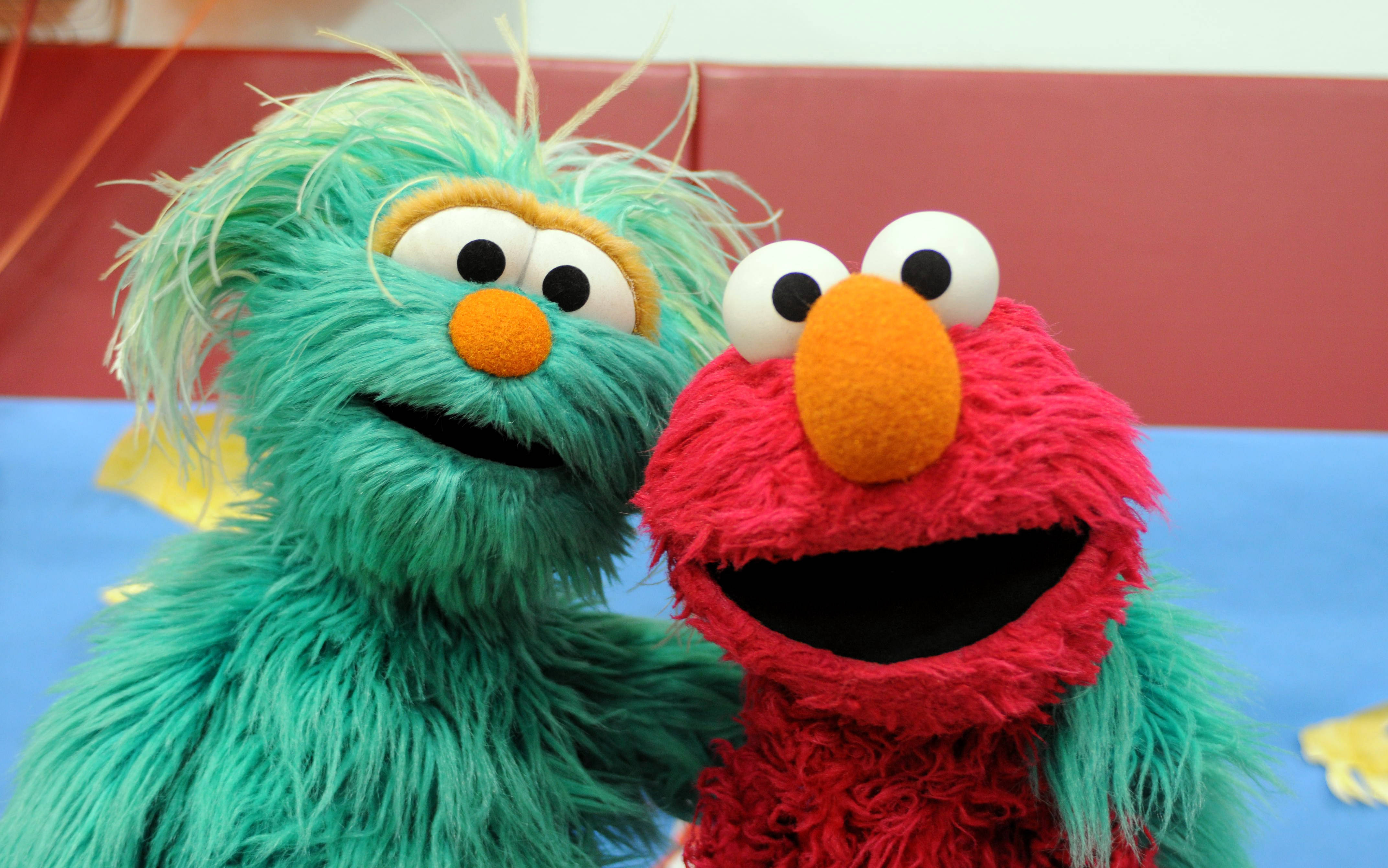 Sesame Street 桌布 壁紙 — 兩個快樂的芝麻街布偶，艾蒙和一個藍綠色毛茸茸的角色，在紅色和藍色背景前燦爛地微笑。