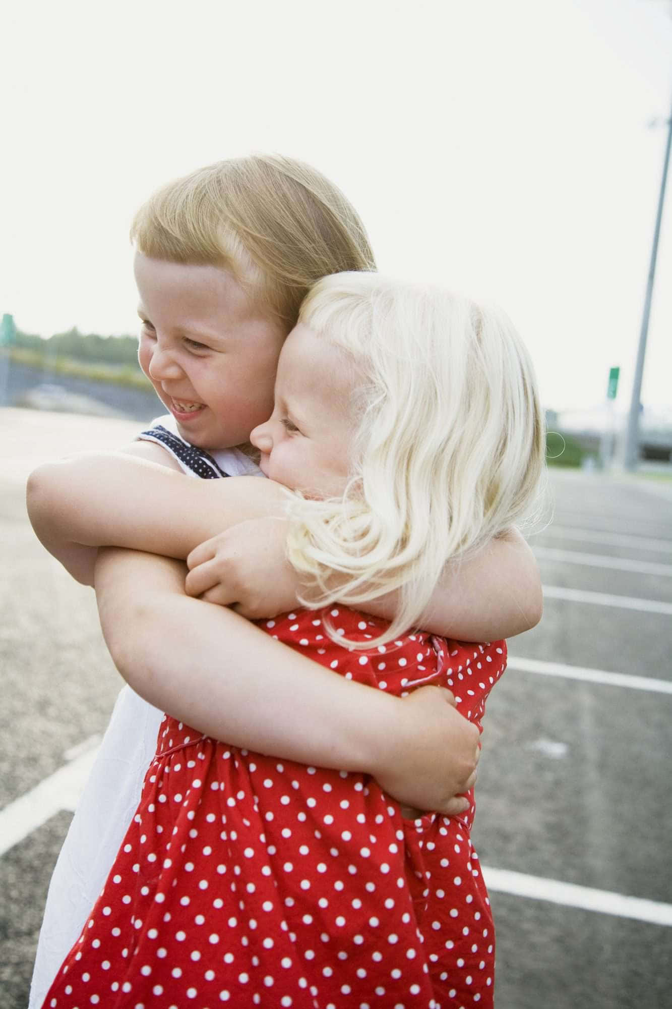Hug 桌布 壁紙 — 兩個快樂的金髮孩子在陽光明媚的停車場熱情擁抱，其中一個穿著紅色圓點連衣裙。