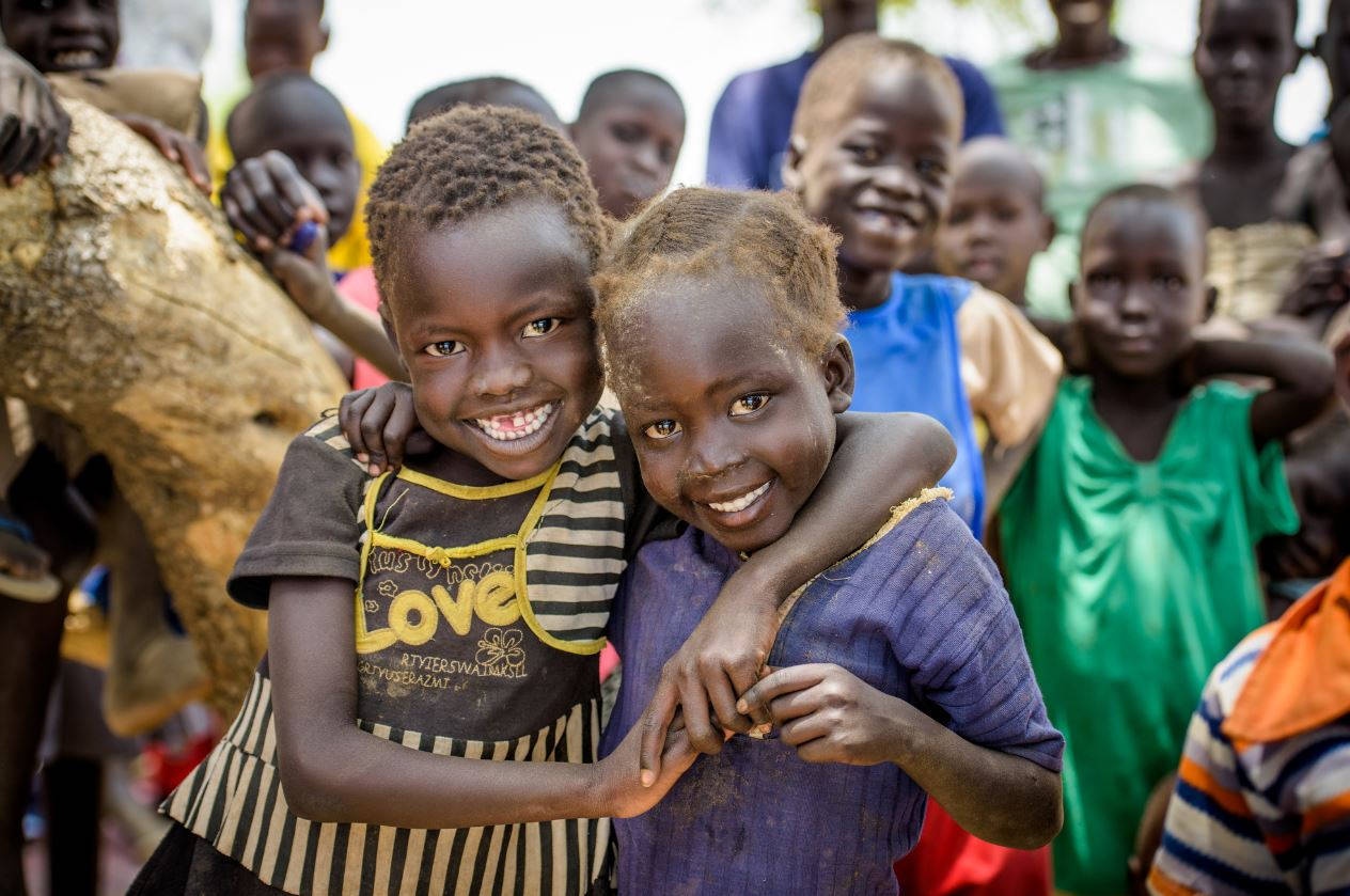 Sudan 桌布 壁紙 — 兩個快樂的非洲兒童帶著燦爛的笑容擁抱，一群同齡人在陽光普照的背景中觀看。