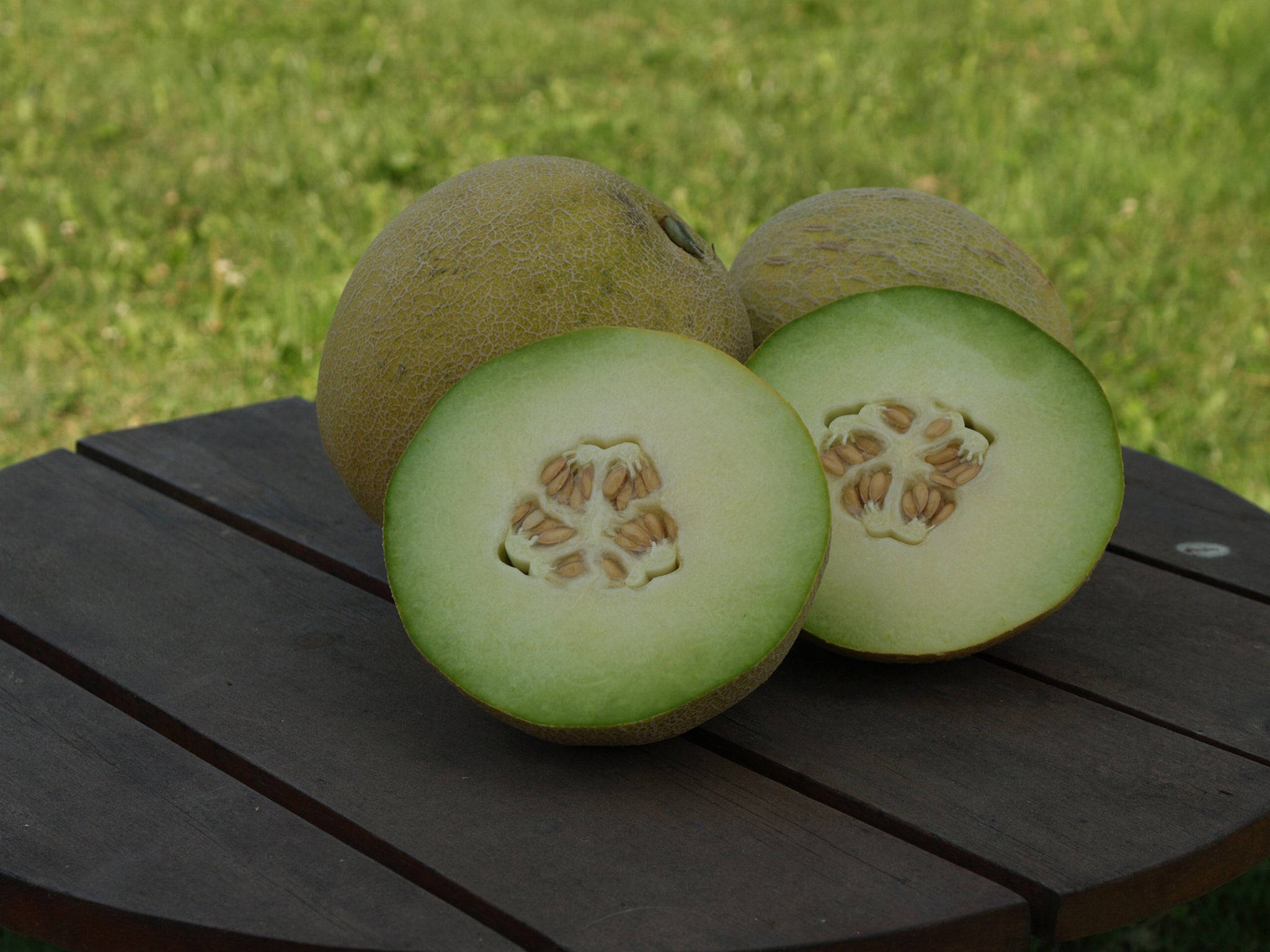Fruits 桌布 壁紙 — 兩個成熟的哈密瓜，其中一個切開，露出淡綠色果肉和種子，放在深色木桌上，背景是模糊的綠色草地。