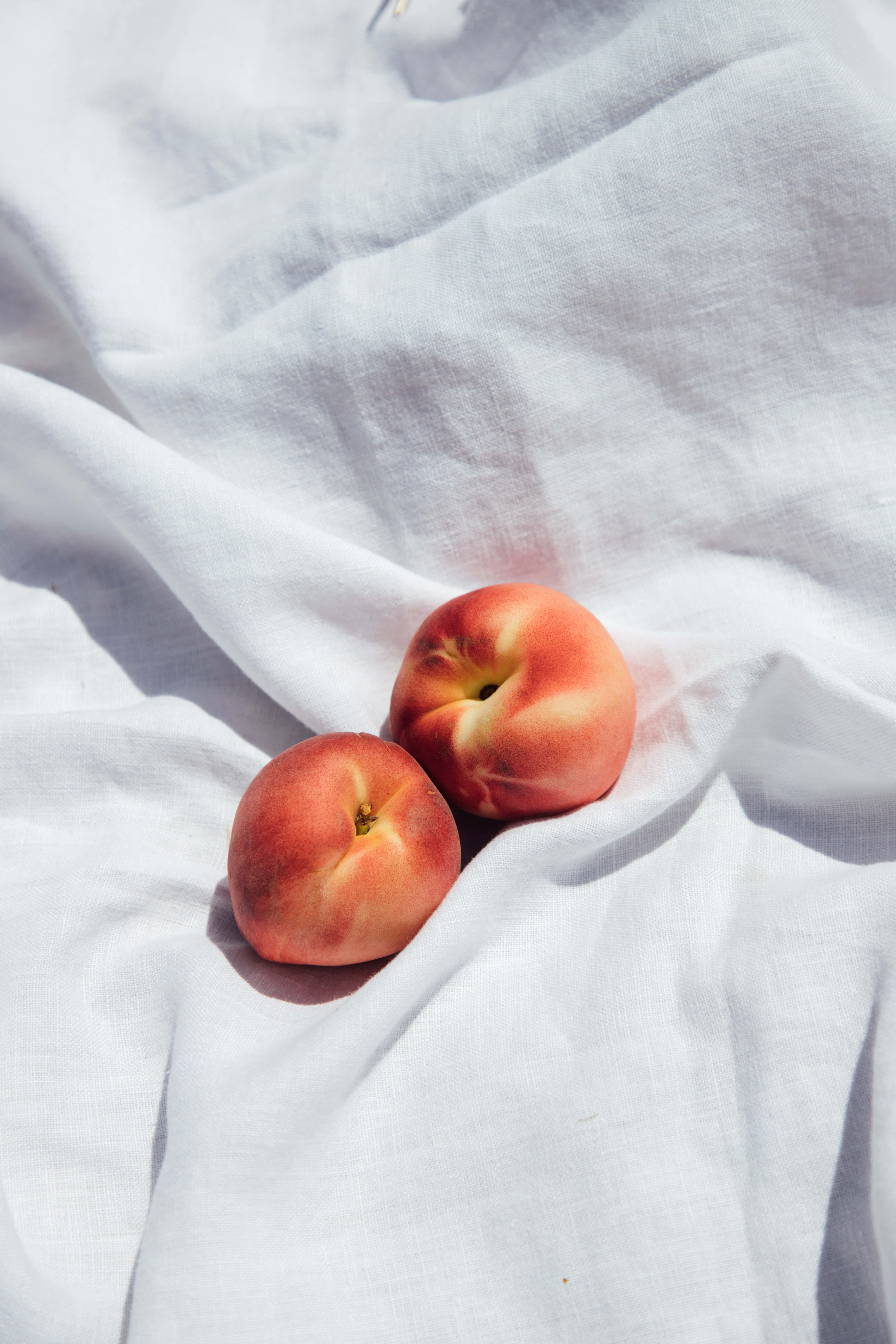 Peaches 桌布 壁紙 — 兩個成熟的桃子在陽光照射的白色亞麻布上，投下柔和的陰影。