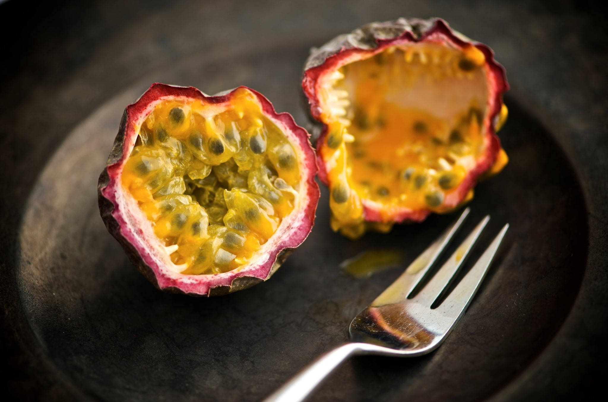 Passion Fruit 桌布 壁紙 — 兩個成熟的百香果半邊，帶有鮮黃色的果肉和黑色的種子，放在一個深色盤子上，旁邊有一把銀叉。