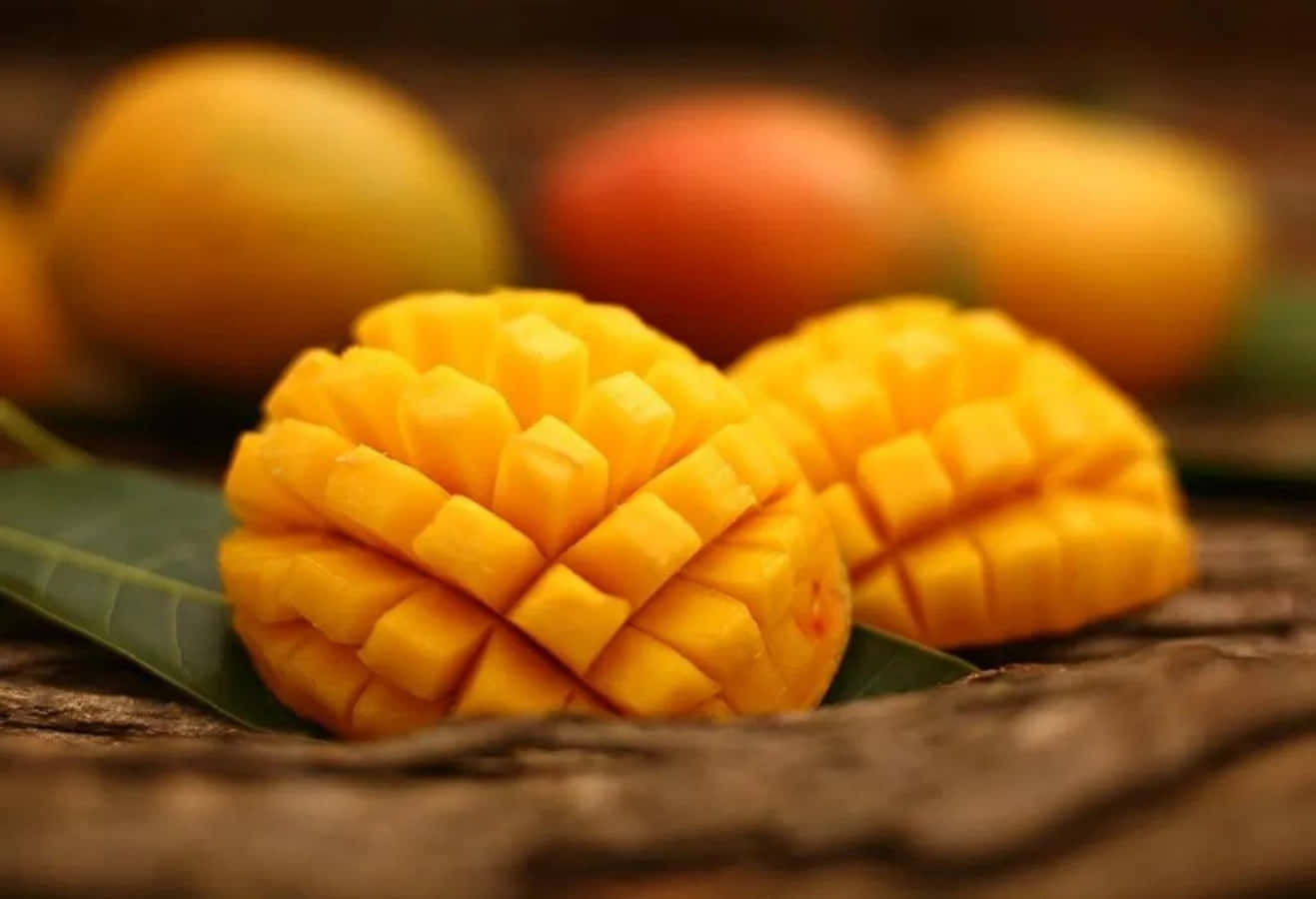 Mango 桌布 壁紙 — 兩個成熟的芒果切成方塊，放在綠葉上，置於質樸的木質表面，背景有模糊的完整芒果。