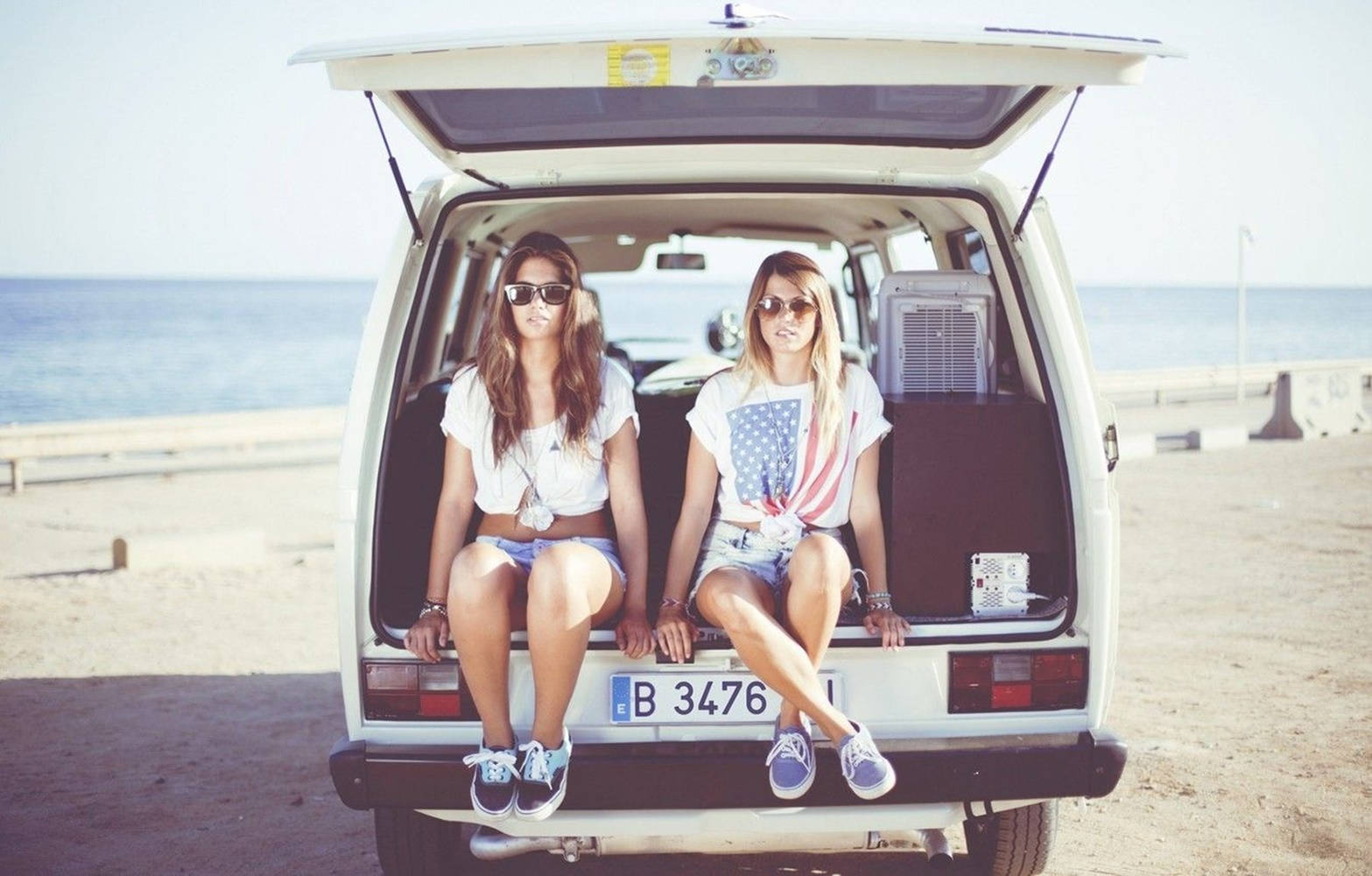 Friends 桌布 壁紙 — 兩個戴太陽眼鏡的女孩隨意地坐在一輛白色貨車的打開尾門上，貨車停泊在陽光明媚的海灘，後面是海洋。