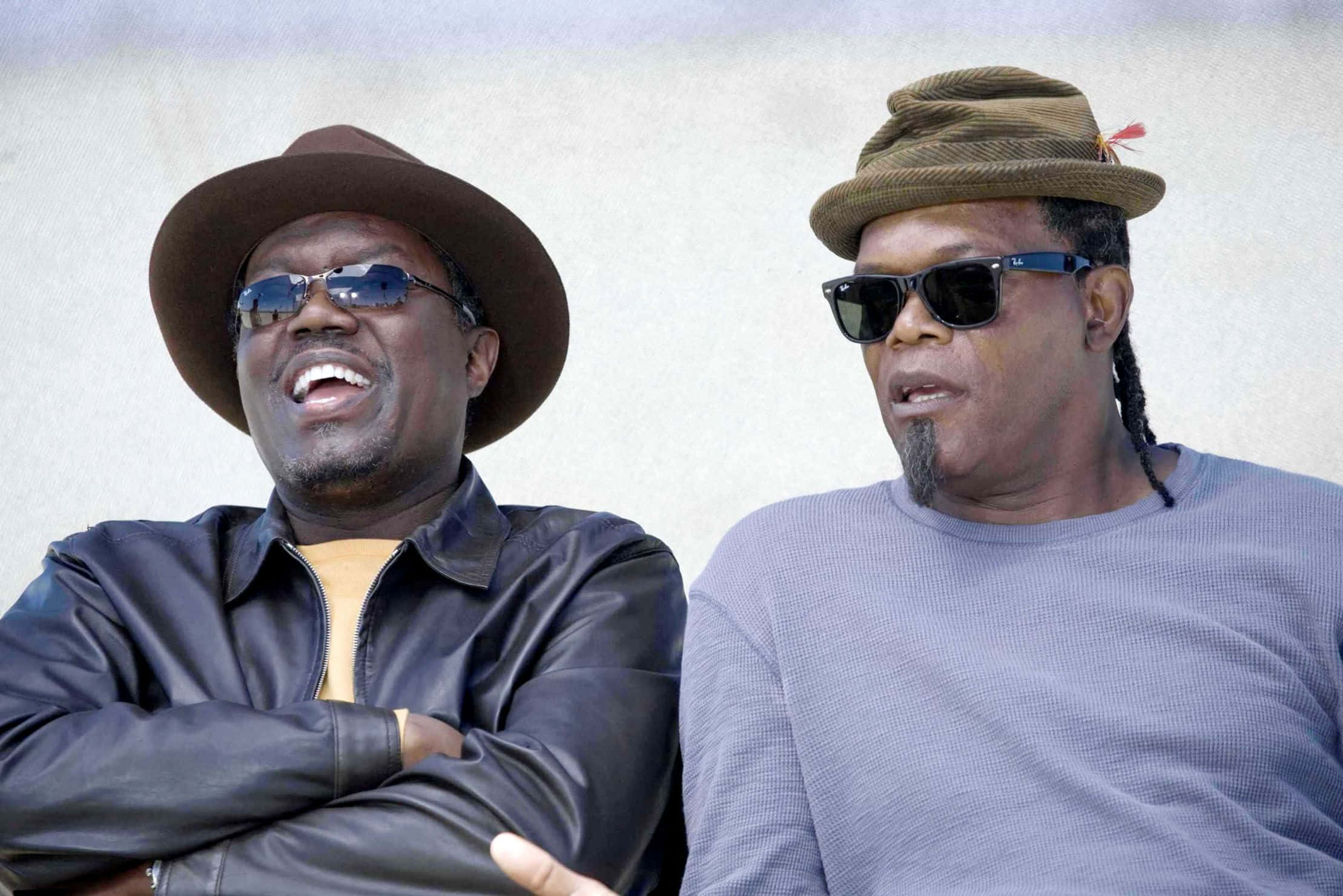 Bernie Mac 桌布 壁紙 — 兩個戴帽子和太陽眼鏡的男人在樸素的紋理牆前一起大笑。