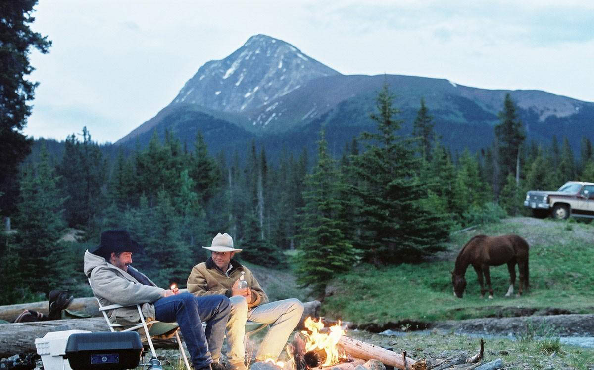 brokeback mountain 桌布 壁紙 — 兩個戴牛仔帽的男人在營火旁放鬆，附近有一匹馬在吃草，背景是高聳的雪山。