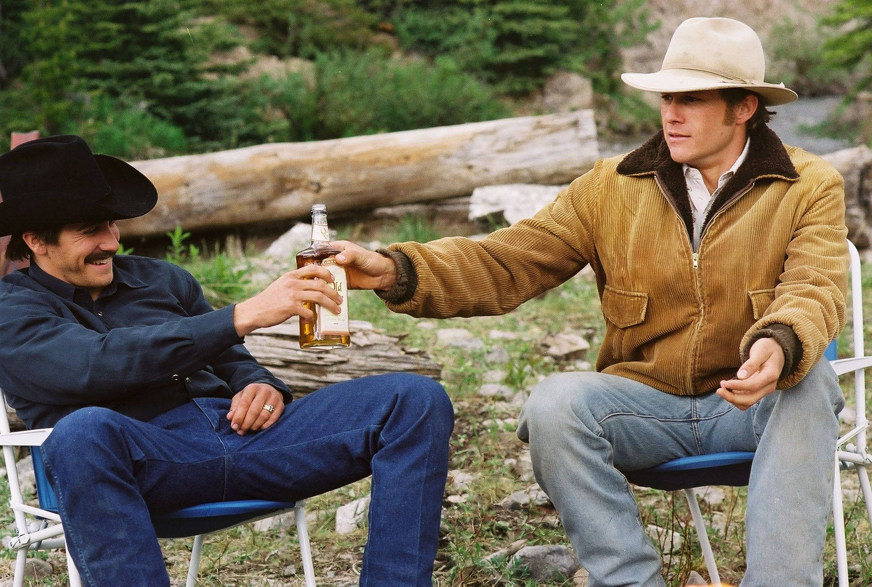 brokeback mountain 桌布 壁紙 — 兩個戴牛仔帽的男人坐在戶外靠近木頭和綠植的地方，用一瓶威士忌乾杯。