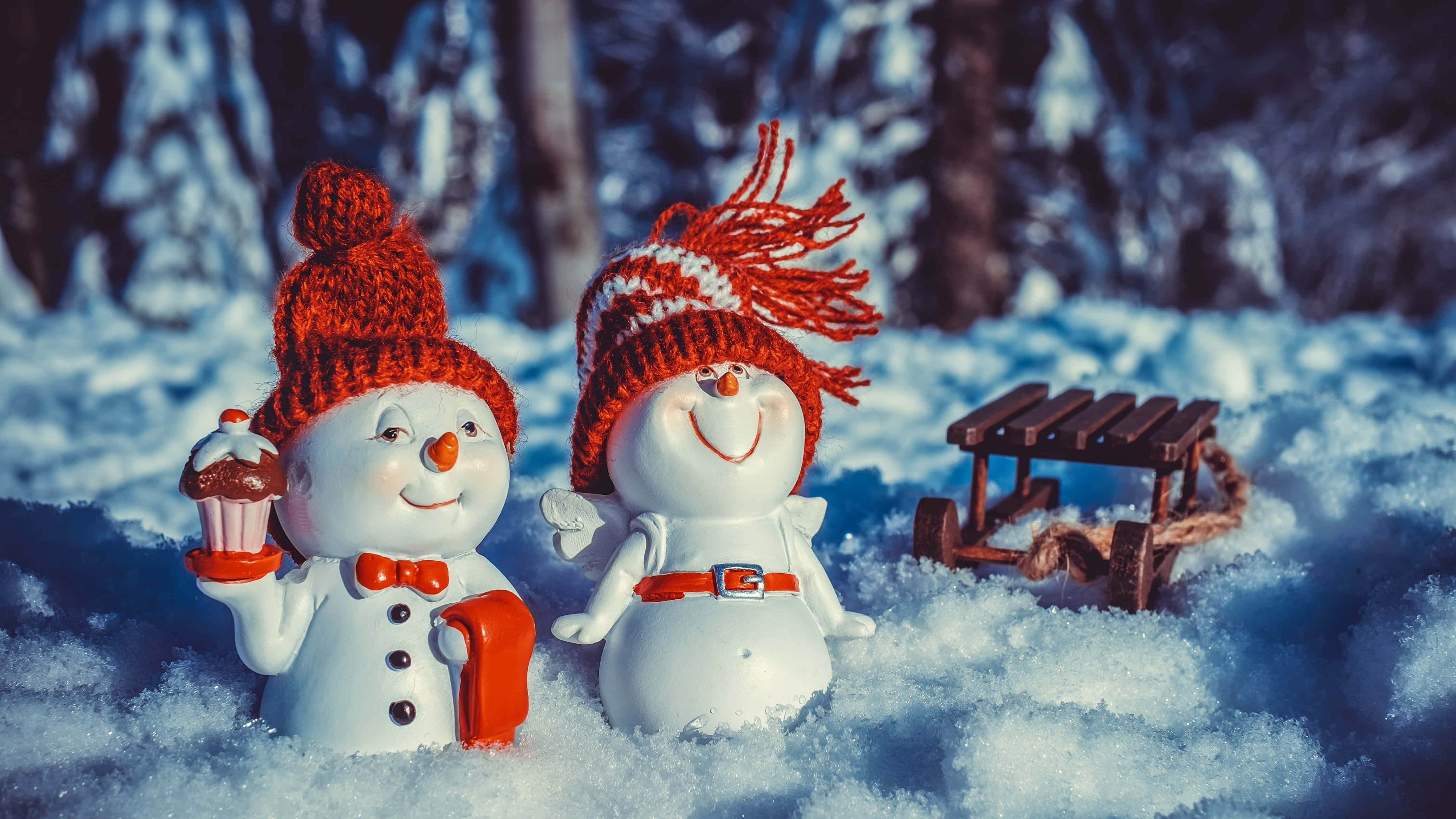 聖誕節 桌布 壁紙 — 兩個戴紅色針織帽的快樂雪人公仔站在深雪中，旁邊有一個木製雪橇，在冬季森林裡。