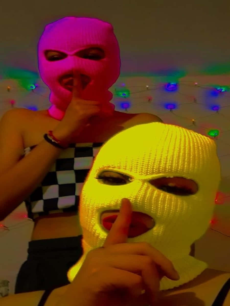 Ski Mask Girl 桌布 壁紙 — 兩個戴著亮粉紅色和黃色巴拉克拉法帽的人在彩色燈串下做出噤聲手勢。