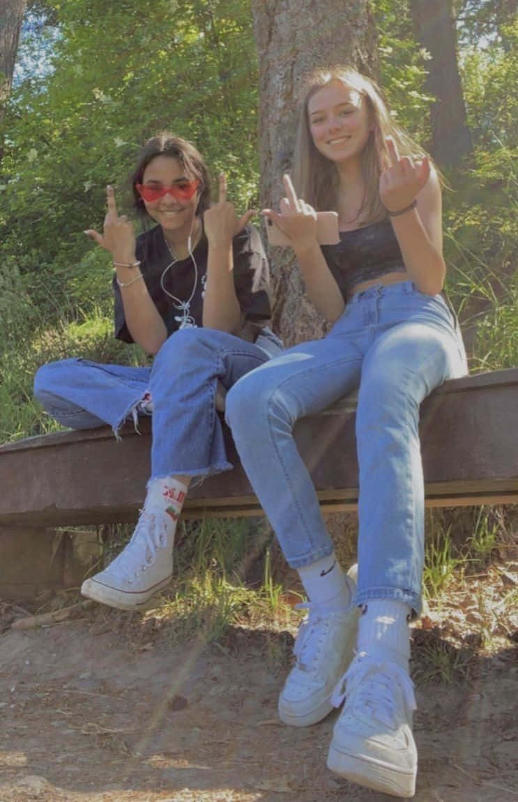 Best Friend Aesthetic 桌布 壁紙 — 兩個戴著太陽眼鏡和牛仔褲的微笑青少年坐在陽光普照的森林木凳上，做出搖滾手勢。