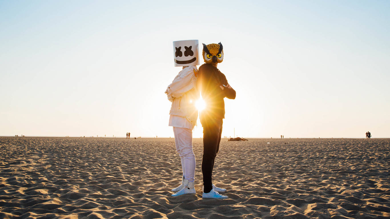 Marshmello 動態圖 桌布 壁紙 — 兩個戴著標誌性面具的人物站在廣闊的沙灘上，背光襯托著金色的日落。