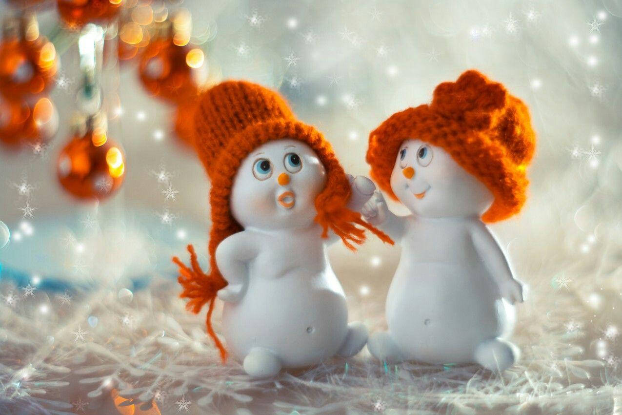 Toy 桌布 壁紙 — 兩個戴著橙色針織帽的可愛雪人公仔一起坐在雪地上，背景有閃爍的節日裝飾和閃閃發光的燈光。