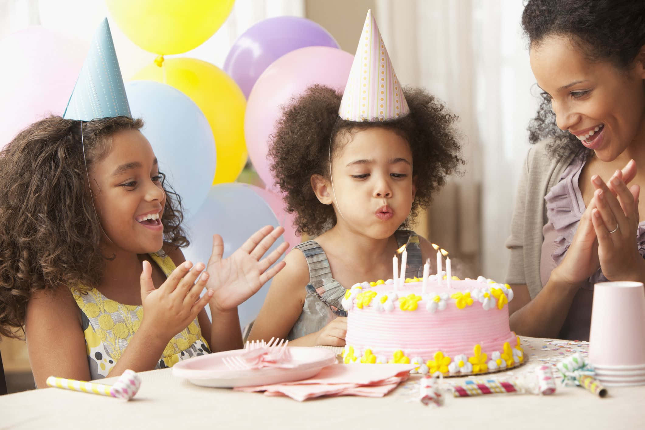 Birthday Party 桌布 壁紙 — 兩個戴著派對帽的女孩吹熄粉紅色生日蛋糕上的蠟燭，一位大人在旁邊高興地拍手。