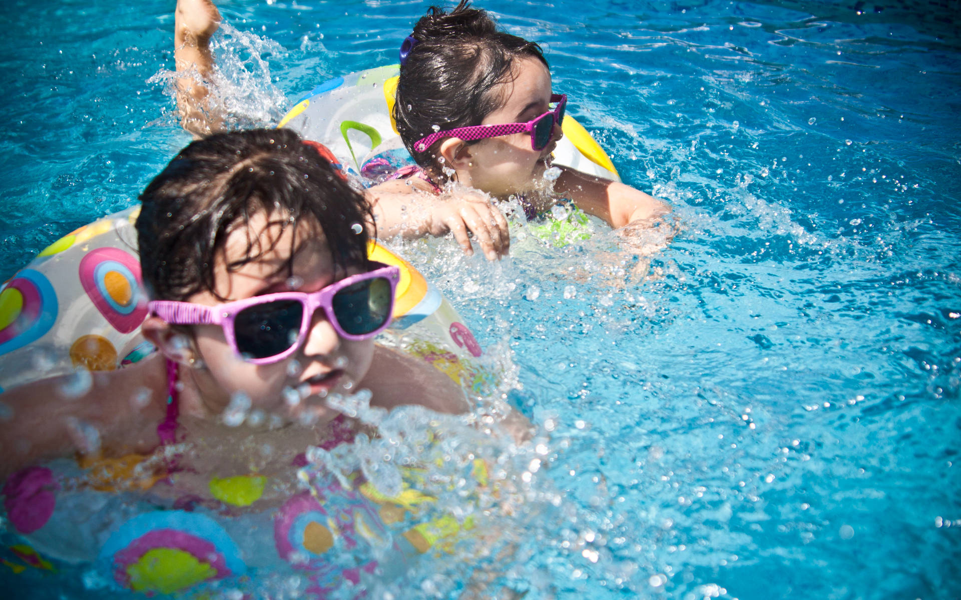 Swimming 桌布 壁紙 — 兩個戴著粉紅色太陽眼鏡的小孩在明亮的藍色泳池中快樂地潑水，漂浮在色彩繽紛的充氣圈上。