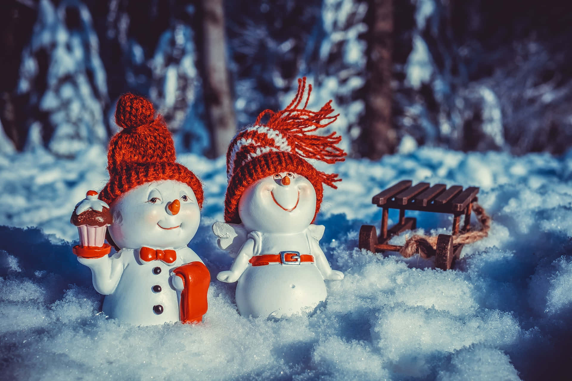 兩個戴紅帽的雪人公仔在雪地森林中拿著一個杯子蛋糕和一個雪橇。 — 雪人 壁紙