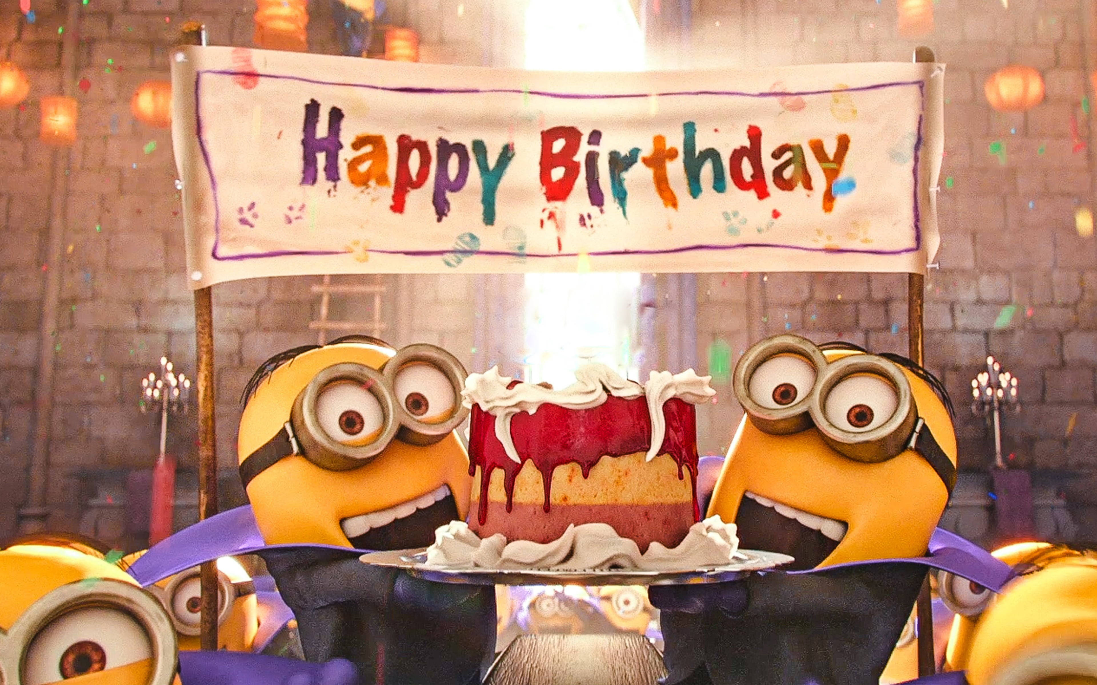 Birthday Cake 桌布 壁紙 — 兩個戴著紫色蝴蝶結的Minions在色彩繽紛的「Happy Birthday」橫幅和飄落的五彩紙屑下，愉快地展示一個紅色生日蛋糕。