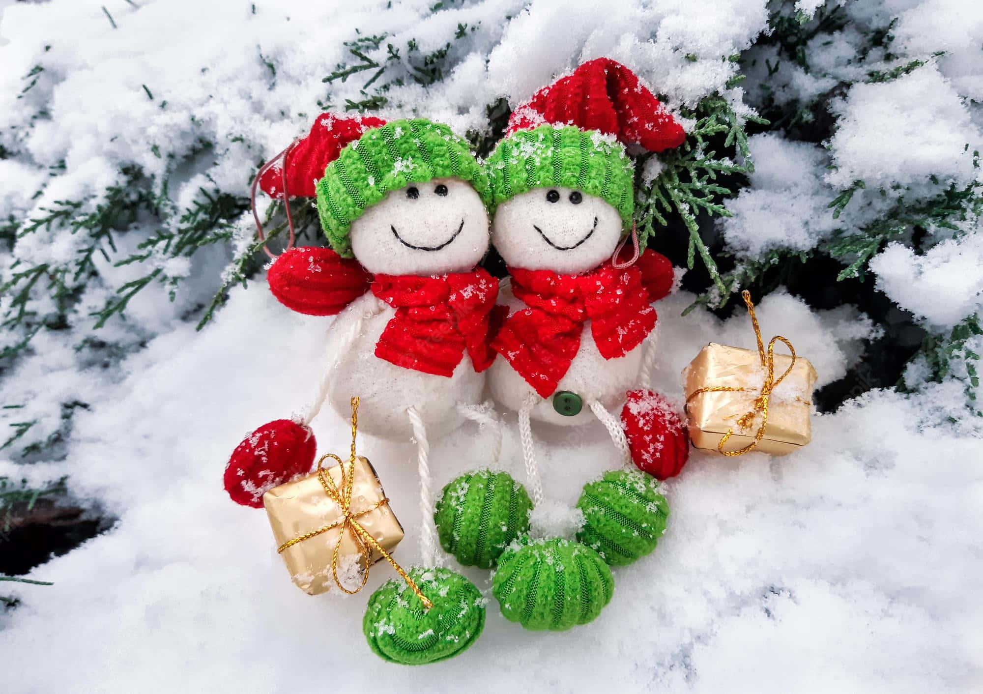 雪 桌布 壁紙 — 兩個戴著綠色帽子和紅色圍巾的快樂雪人裝飾品坐在白雪覆蓋的松樹枝上，旁邊是金色禮物和綠色聖誕球。