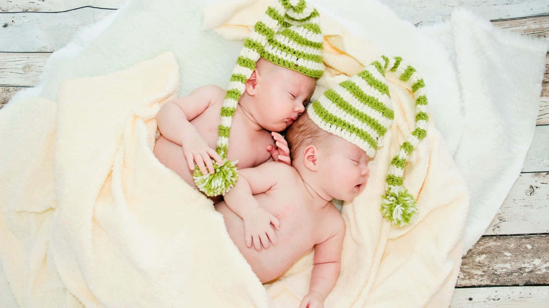 twins 桌布 壁紙 — 兩個戴著綠色條紋針織帽的新生雙胞胎在破舊木地板上柔軟的黃色毯子上安詳地睡覺。