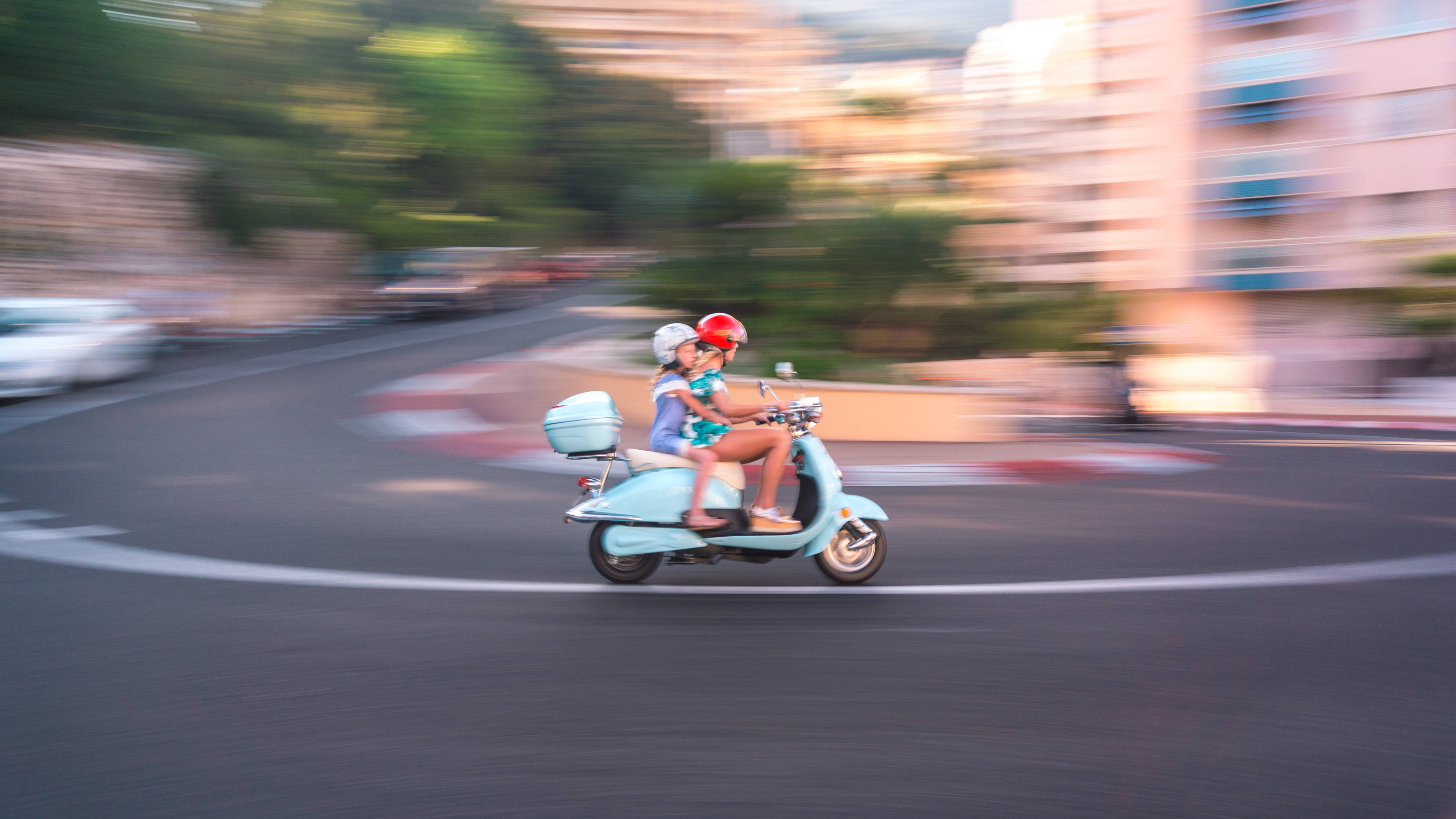 Monaco 桌布 壁紙 — 兩個戴頭盔的孩子在黃金時段騎著淺藍色滑板車穿過模糊的城市街道。
