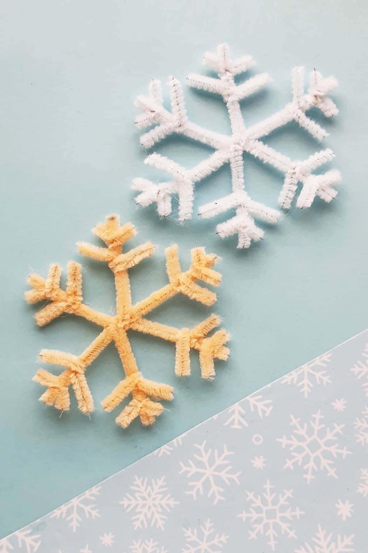 雪花 桌布 壁紙 — 兩個手工製作的管道清潔器雪花——一個白色和一個淡黃色——放在柔軟的藍色表面上，旁邊是一張帶有雪花圖案的紙。