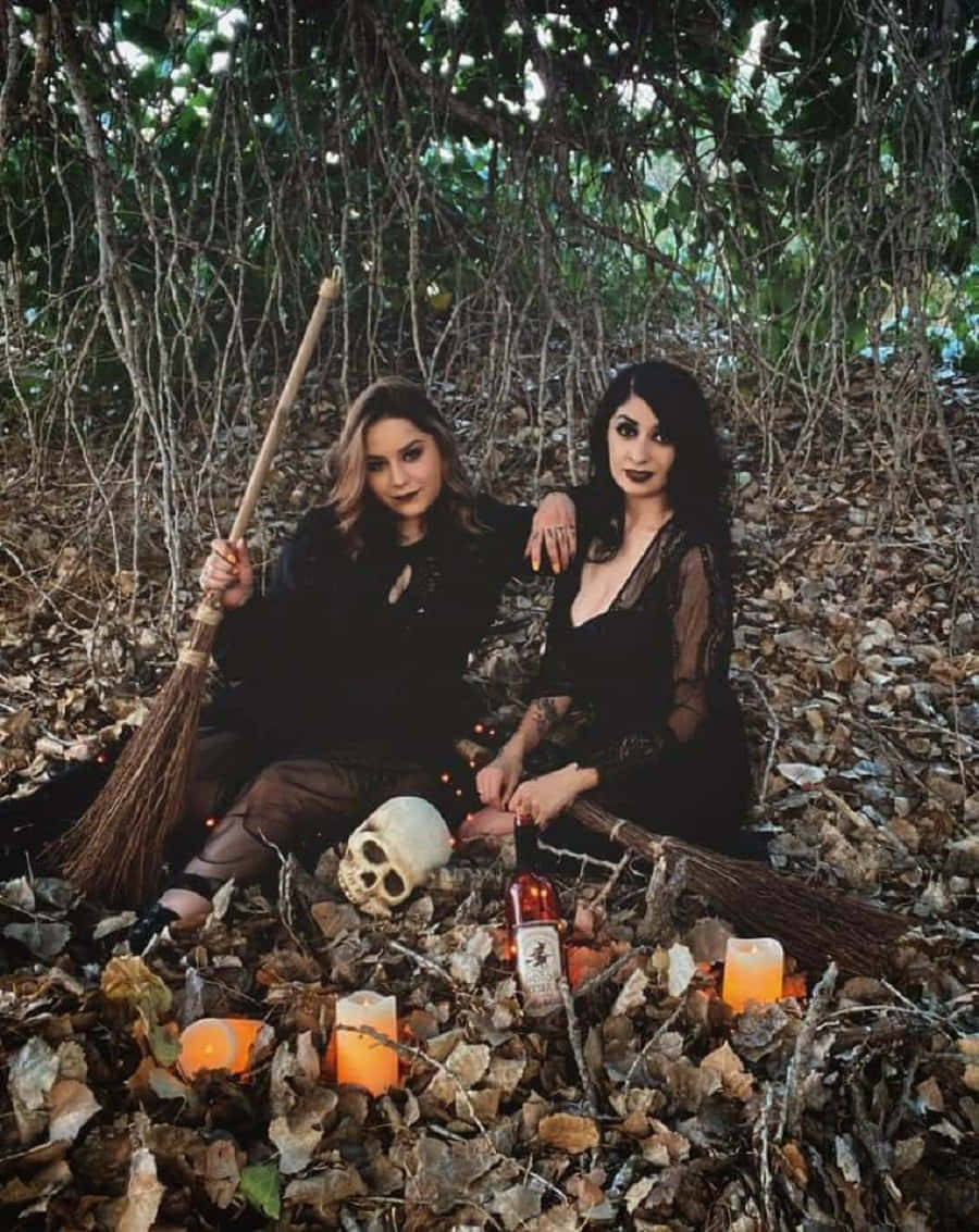 Witch Costumes 桌布 壁紙 — 兩個打扮成女巫的女人坐在落葉中，拿著掃帚，周圍有蠟燭、頭骨和一個瓶子，身處森林中。