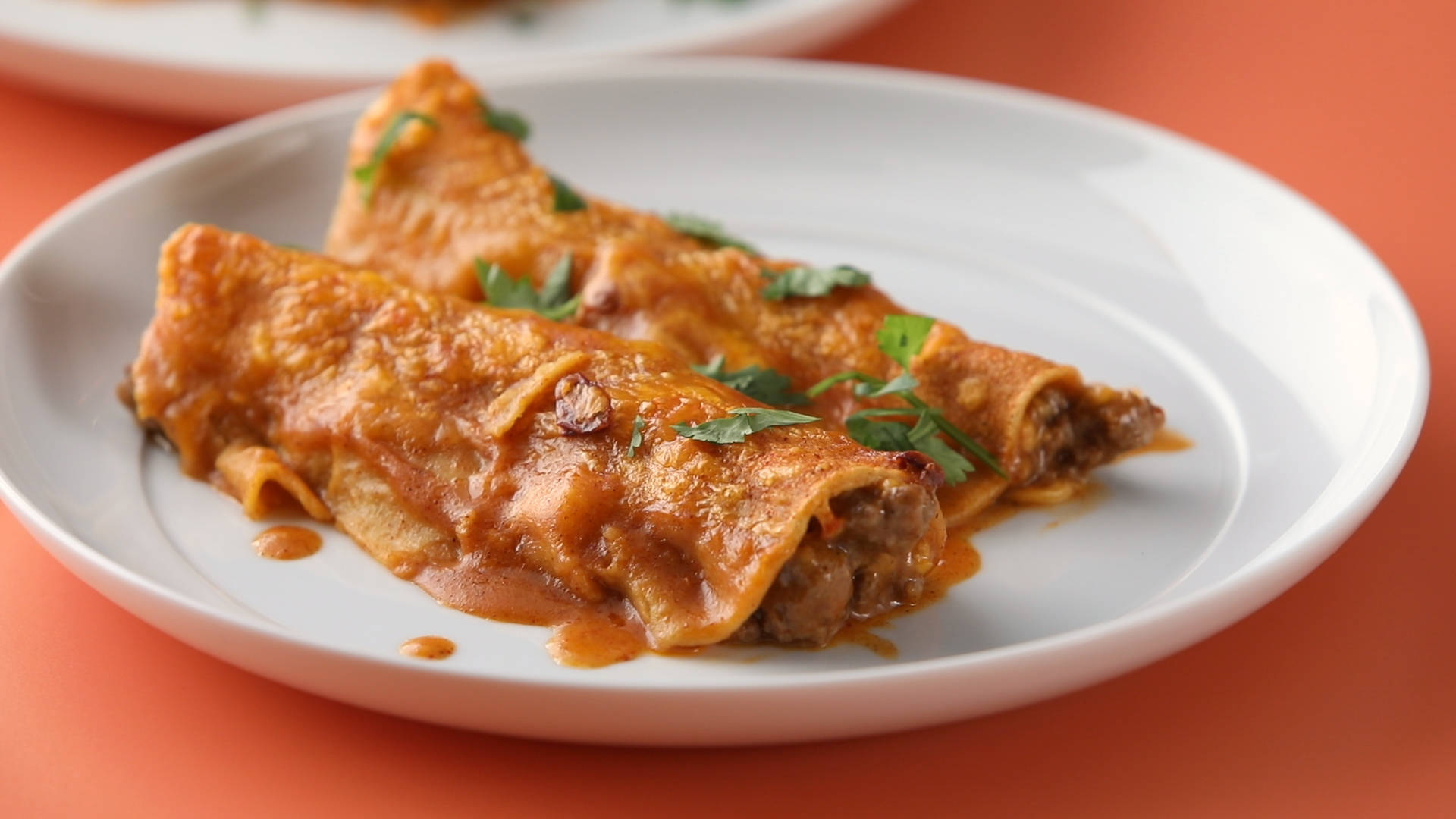 Mexican Food 桌布 壁紙 — 兩個捲好的墨西哥捲餅，淋上濃郁的紅醬和新鮮香菜，放在白色盤子上，背景是溫暖的橙色。