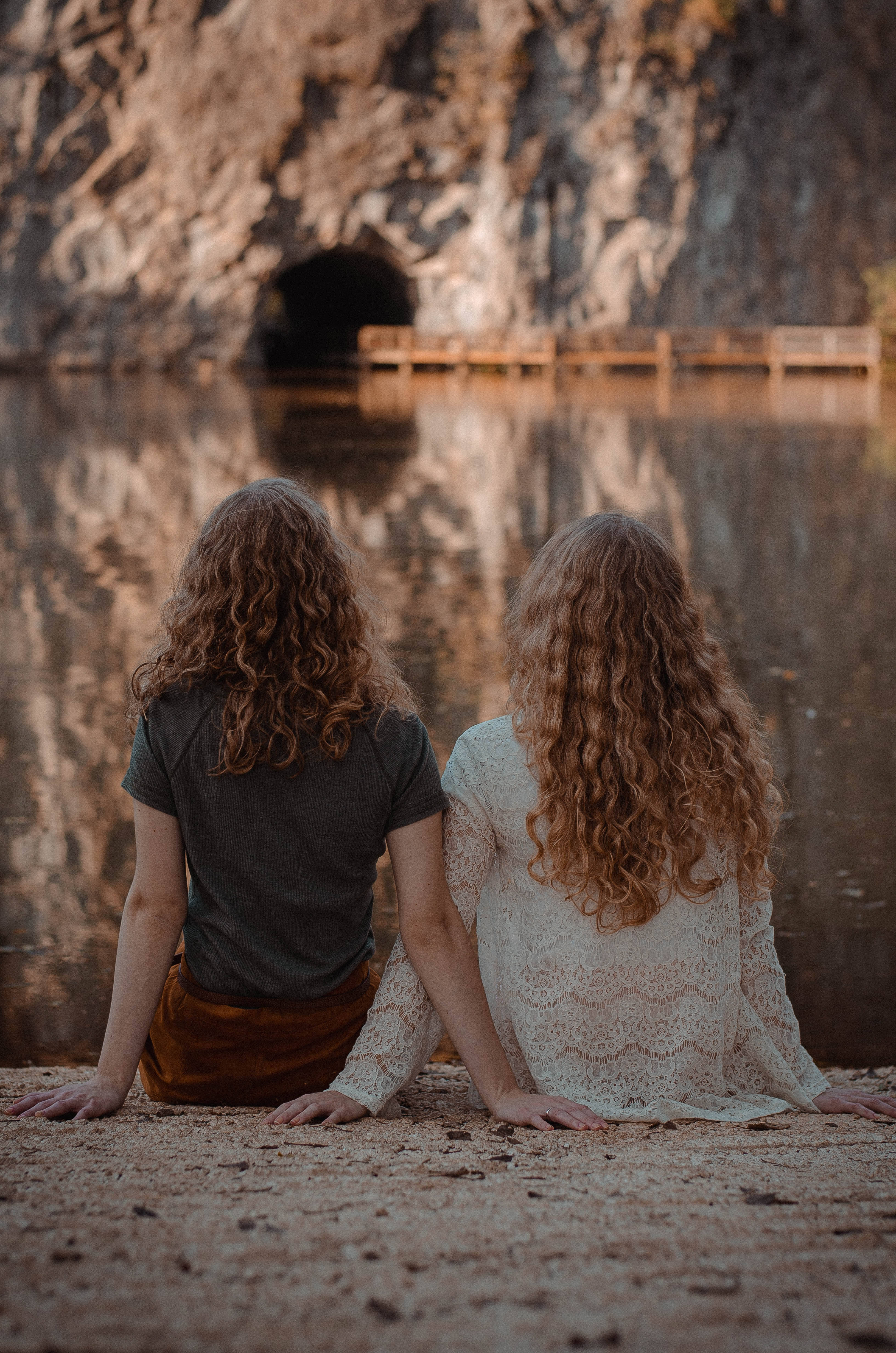 Curly Hair 桌布 壁紙 — 兩個捲髮女孩並肩坐在岩石海岸上，凝視著平靜的湖面，湖面倒映著洞穴入口和木製碼頭。