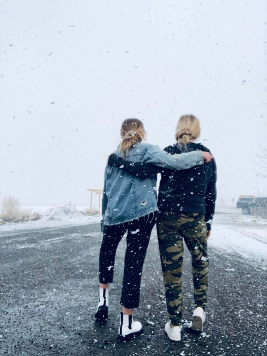 摯友 桌布 壁紙 — 兩個朋友在雪路上從後擁抱，雪花在他們周圍飄落，構成寧靜的冬日場景。