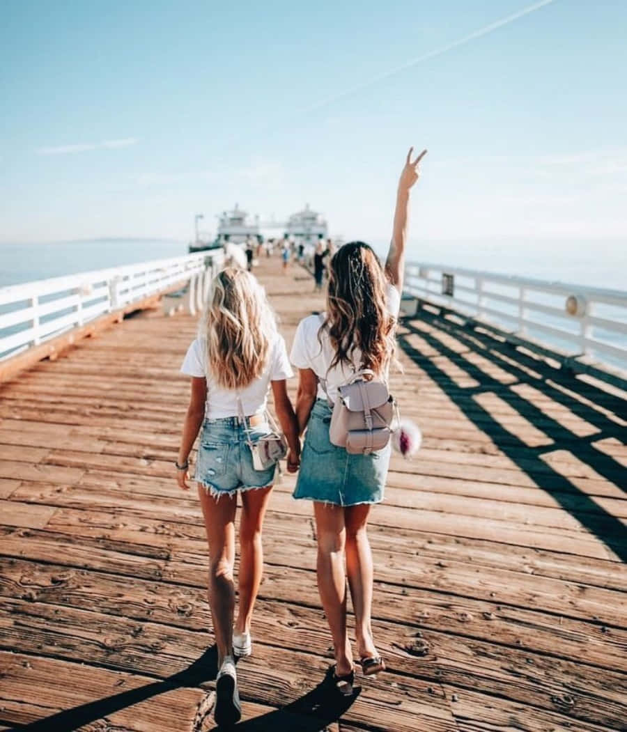 Friends 桌布 壁紙 — 兩個朋友手牽手走在陽光普照的木碼頭上，其中一人愉快地向地平線揮手。