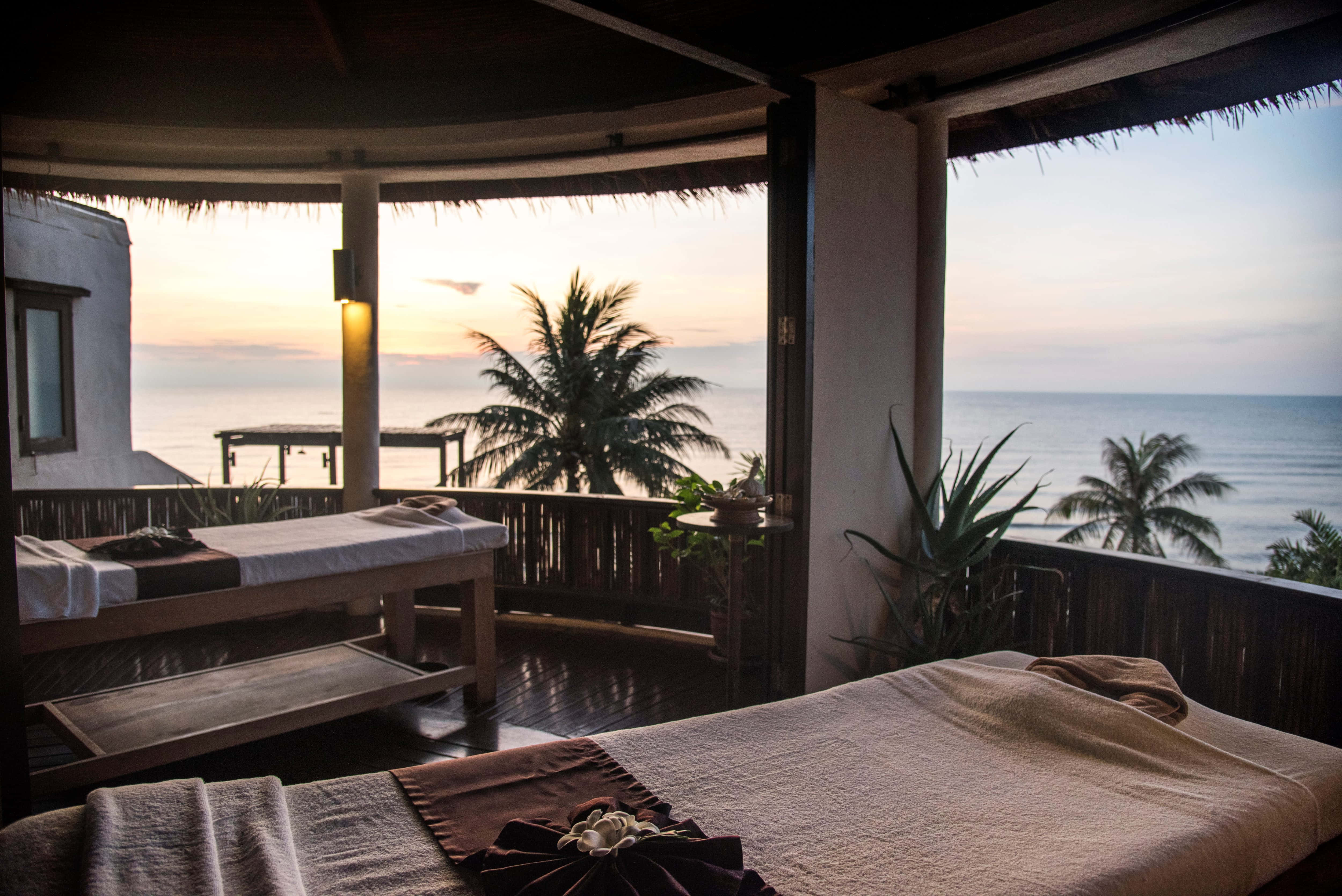 Beach Daybed 桌布 壁紙 — 兩個木製按摩床在茅草屋頂陽台上，俯瞰著棕櫚樹環繞的寧靜海洋日落。