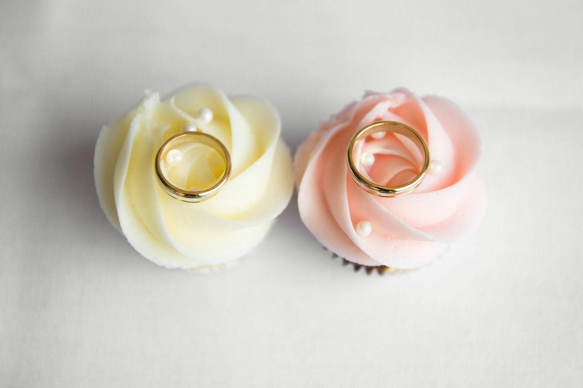 Wedding Rings 桌布 壁紙 — 兩個杯子蛋糕，上面有白色和粉紅色的糖霜，頂部裝飾著金戒指和珍珠，放在純白色的表面上。