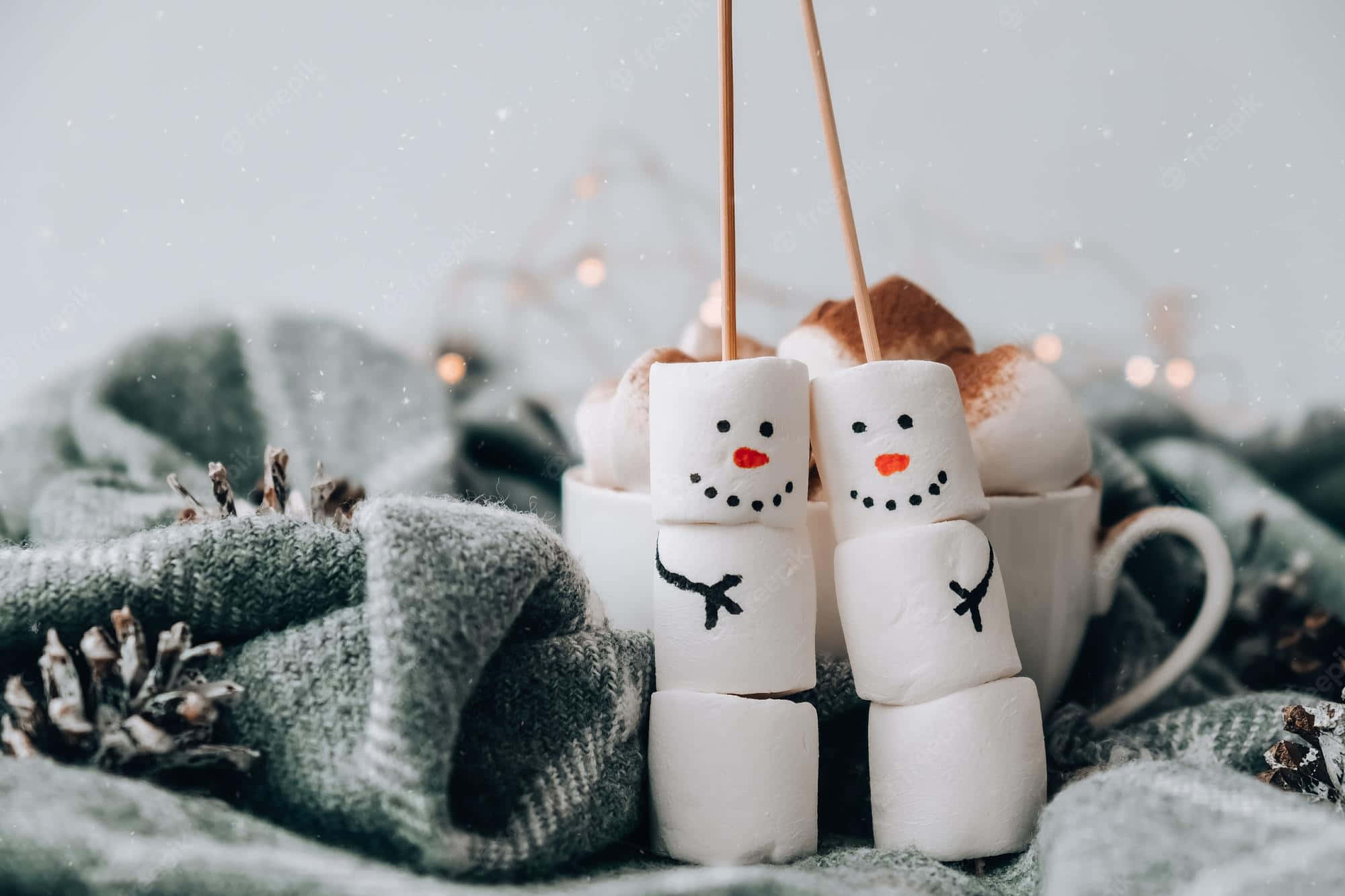 Funny Holiday Desktop 桌布 壁紙 — 兩個棉花糖雪人，有樹枝手臂和胡蘿蔔鼻子，坐在一杯熱可可旁邊，放在舒適的灰色針織毯上。