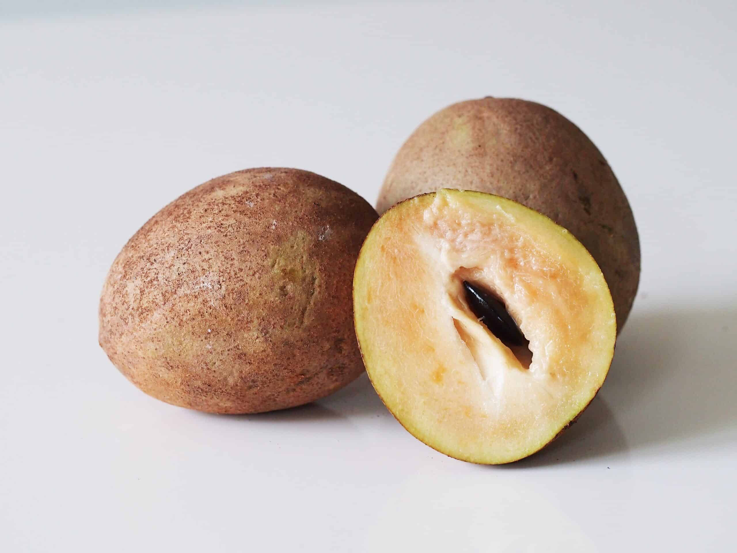 Fruits 桌布 壁紙 — 兩個棕色外皮的沙巴果，其中一個切開，露出奶油黃色的果肉和一顆深色種子，背景為純白色。