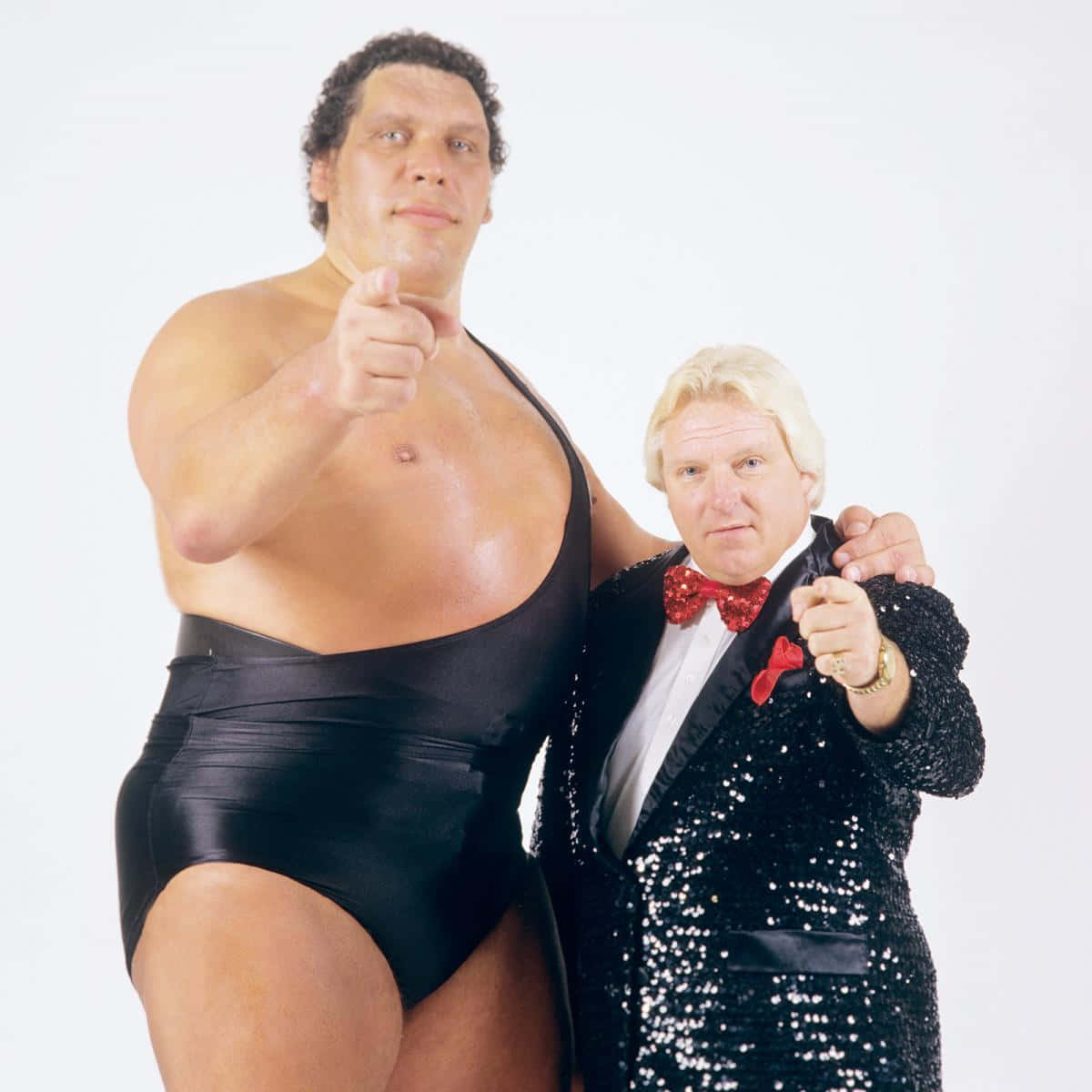 Bobby Heenan 桌布 壁紙 — 兩個標誌性的摔角人物在純白色背景前自信地擺姿勢，一個穿著黑色摔角緊身衣，另一個穿著閃亮的黑色燕尾服配紅色領結，兩人都指向前方。