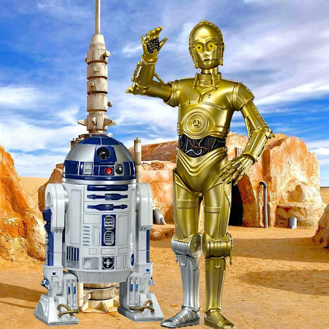 Tatooine 桌布 壁紙 — 兩個標誌性的星際大戰機器人R2-D2和C-3PO站在沙漠峽谷中，頭頂是明亮的藍天，散佈著幾朵雲。