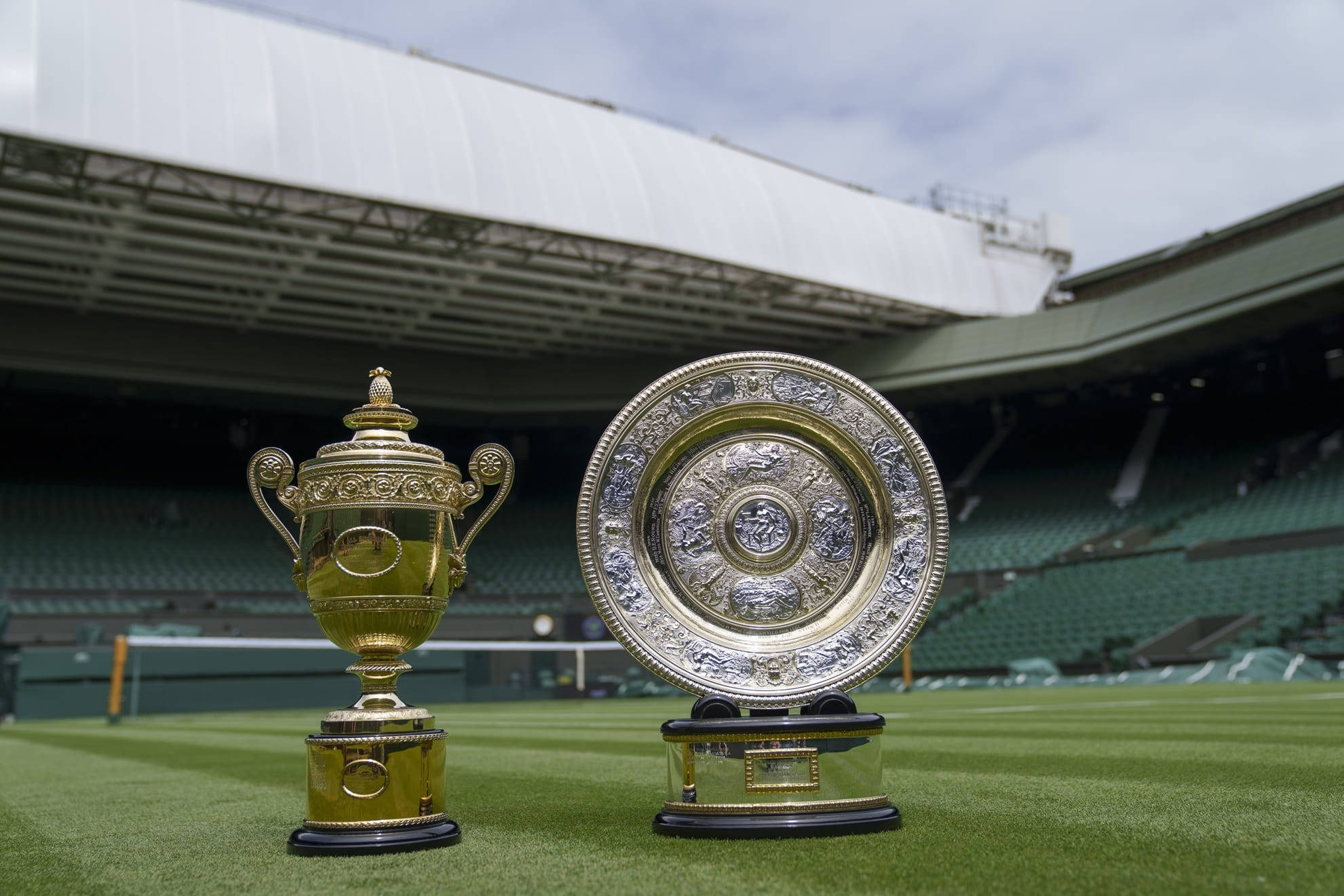 Wimbledon 桌布 壁紙 — 兩個標誌性網球獎盃在草地上閃耀，背景是陰天和空無一人的體育場座位。