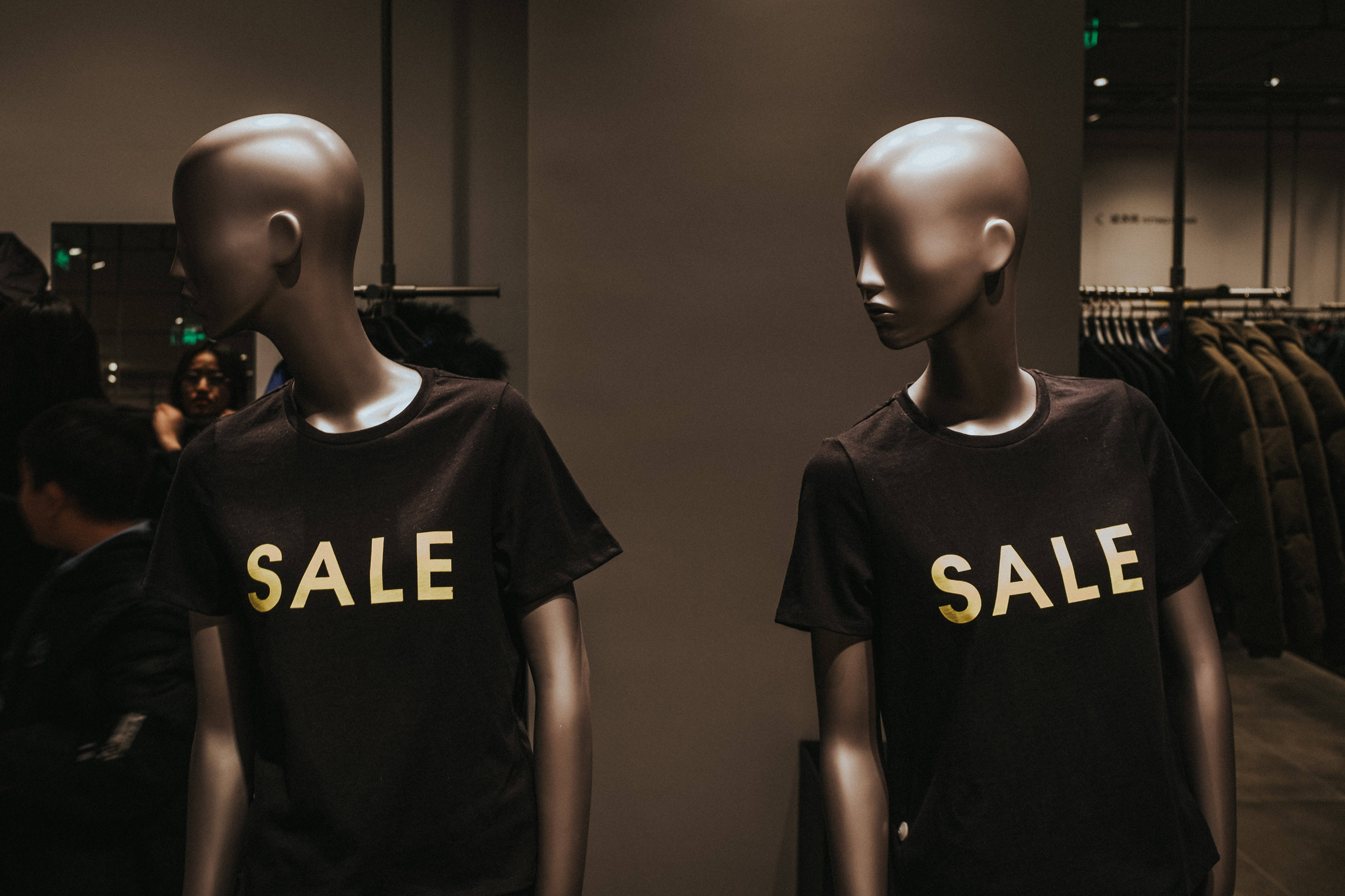 Sales 桌布 壁紙 — 兩個模特兒在燈光昏暗的商店裡穿著黑色T恤，上面有粗體金色「SALE」字樣，彼此面對面。