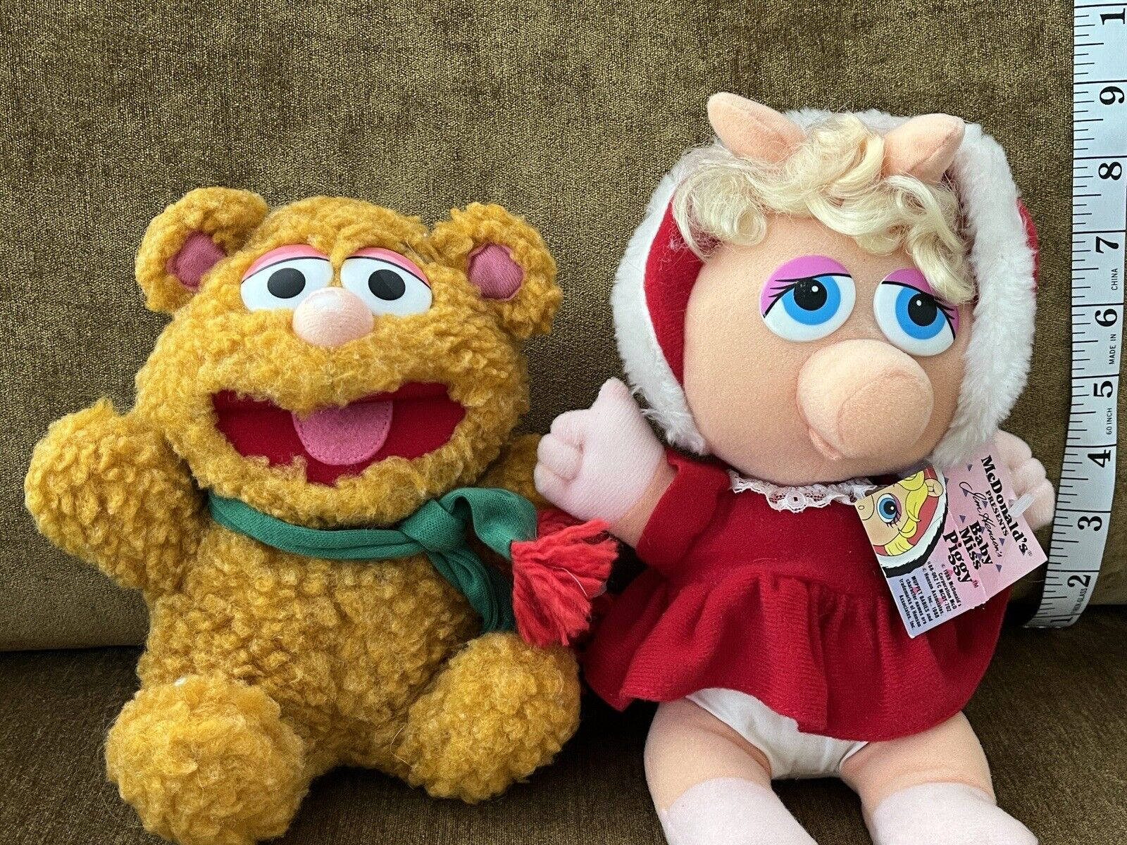 Fozzie Bear 桌布 壁紙 — 兩個毛絨Muppet公仔，一隻戴綠色圍巾的黃色熊和一隻穿著紅色冬季服裝的豬，並排坐在棕色沙發上，旁邊有一個軟尺。