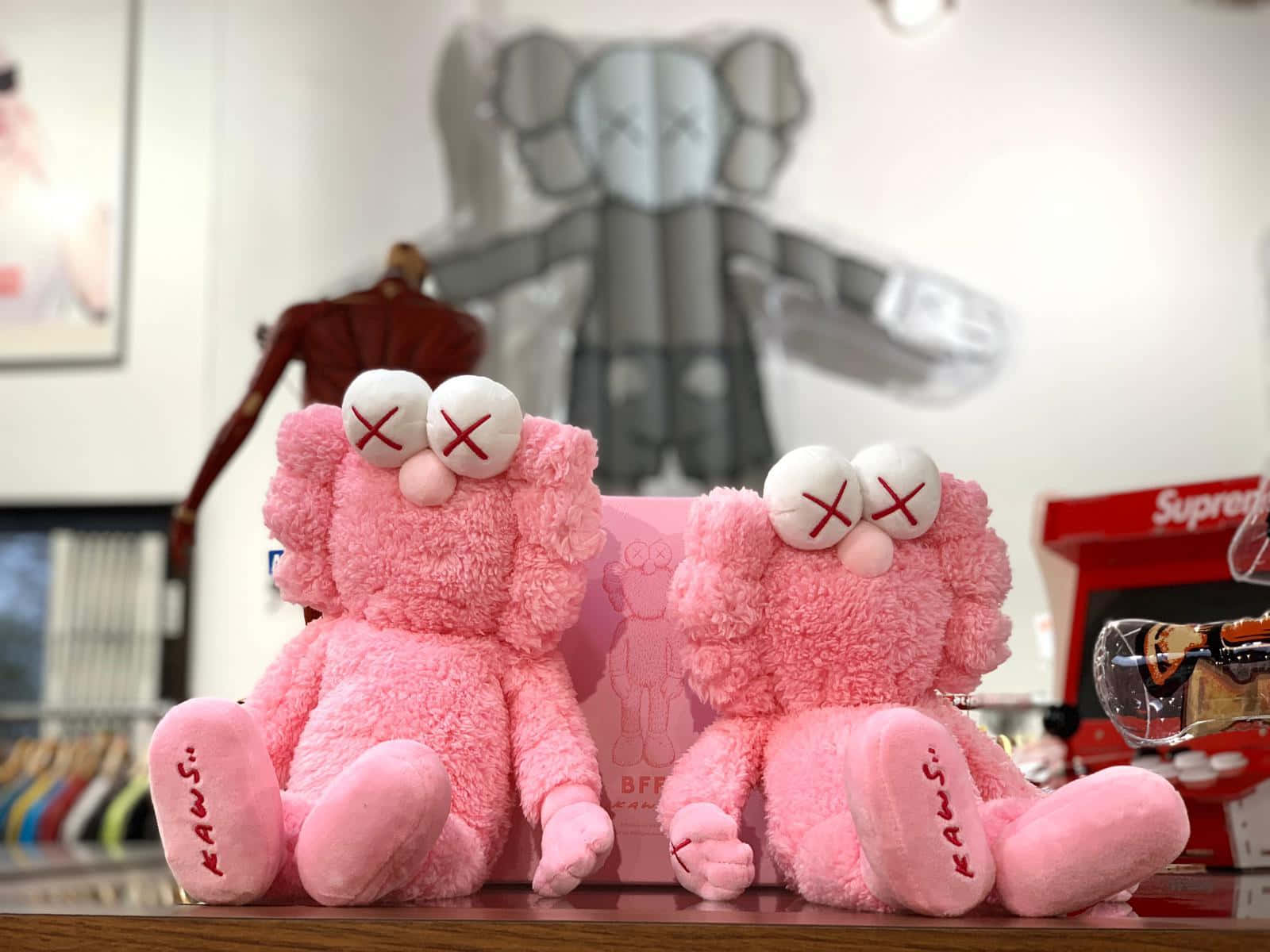 Kaws Bff Pink 桌布 壁紙 — 兩個毛茸茸的粉紅色KAWS毛絨玩具並排坐在木製櫃檯上，背景有一個灰色KAWS雕塑。